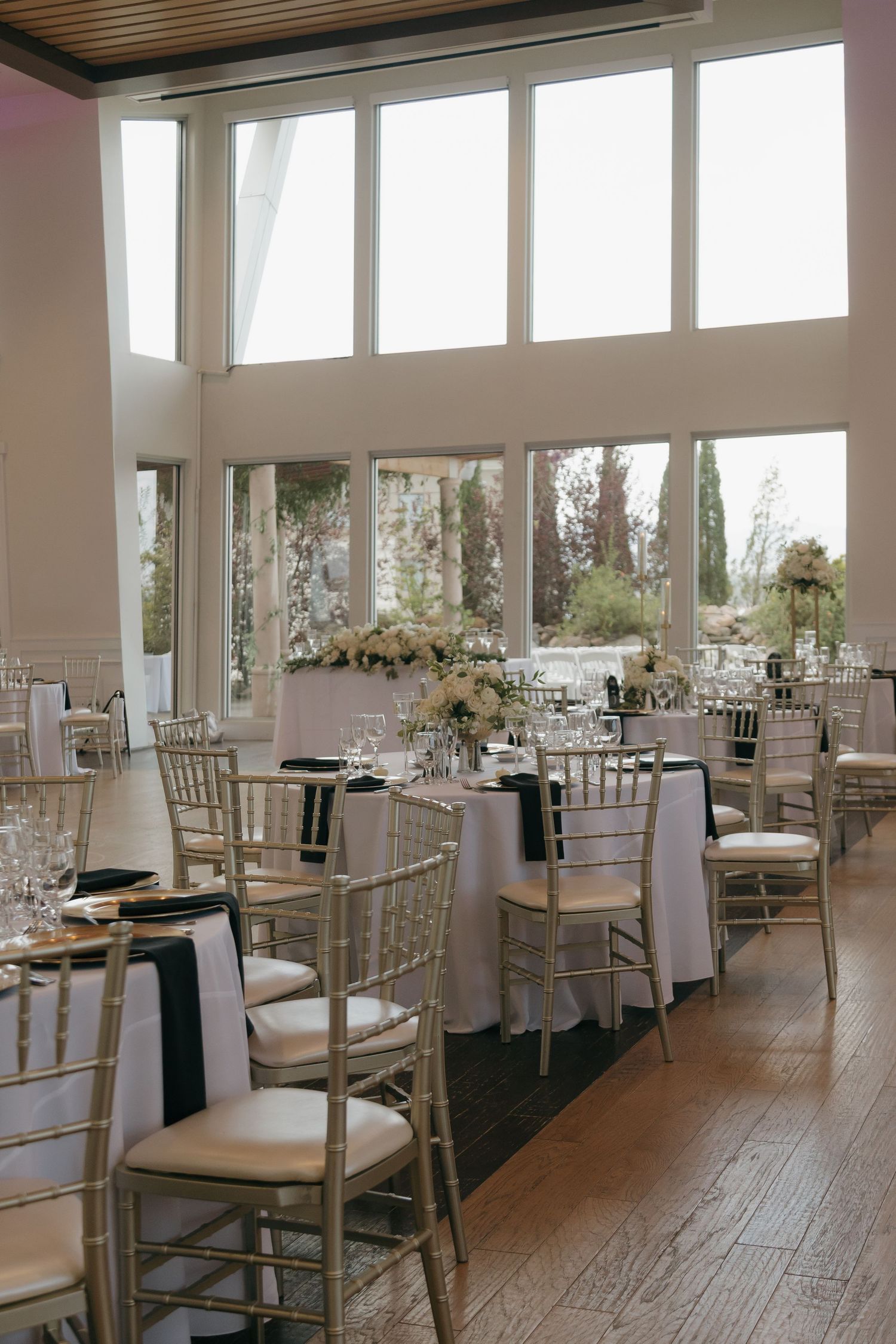 Elegant wedding reception hall with gold chiavari chairs, white floral centerpieces, and tall windows.