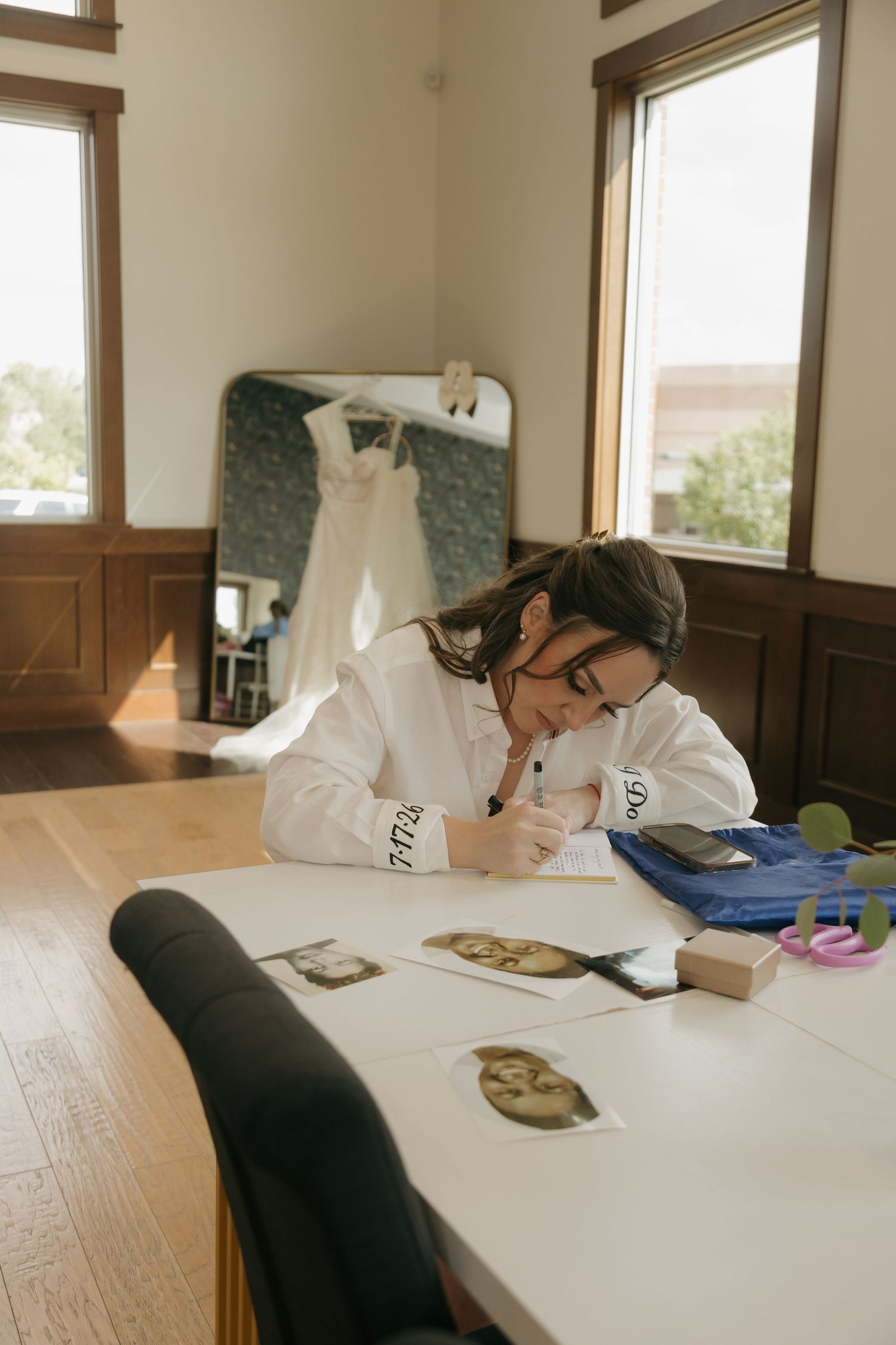 Bride writing notes at a wooden table with wedding dress hanging in background mirror reflection.