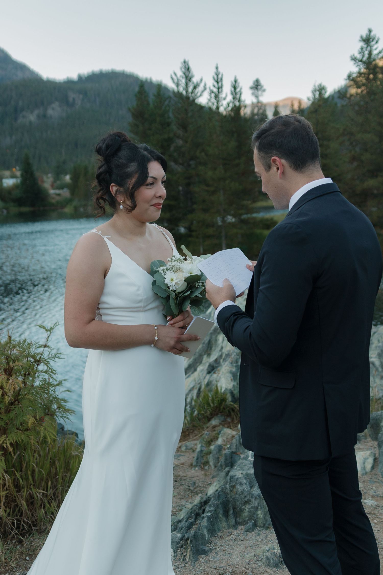 A scenic lakeside wedding ceremony with mountains and evergreen trees in the background.