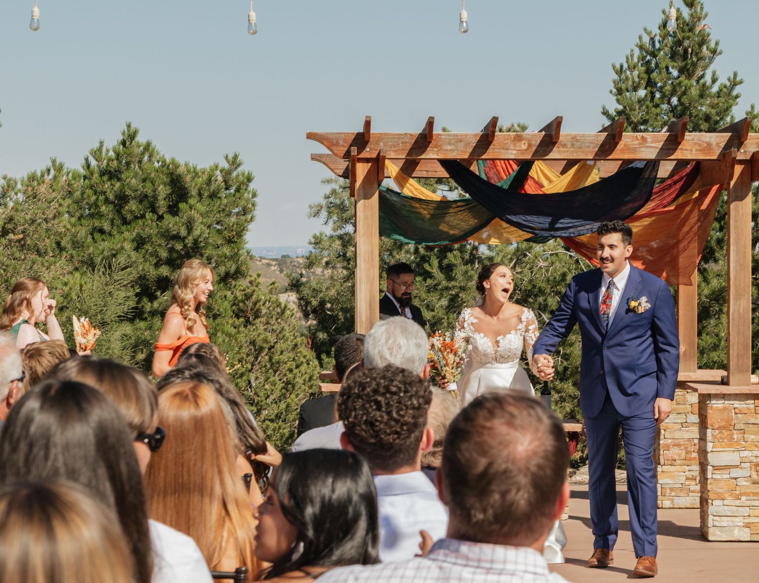 Outdoor wedding ceremony with guests seated beneath a wooden pergola against mountain backdrop.