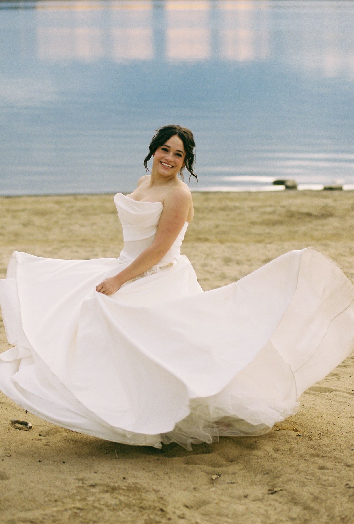 A white wedding dress twirls gracefully on a beach with ocean waves in the background.