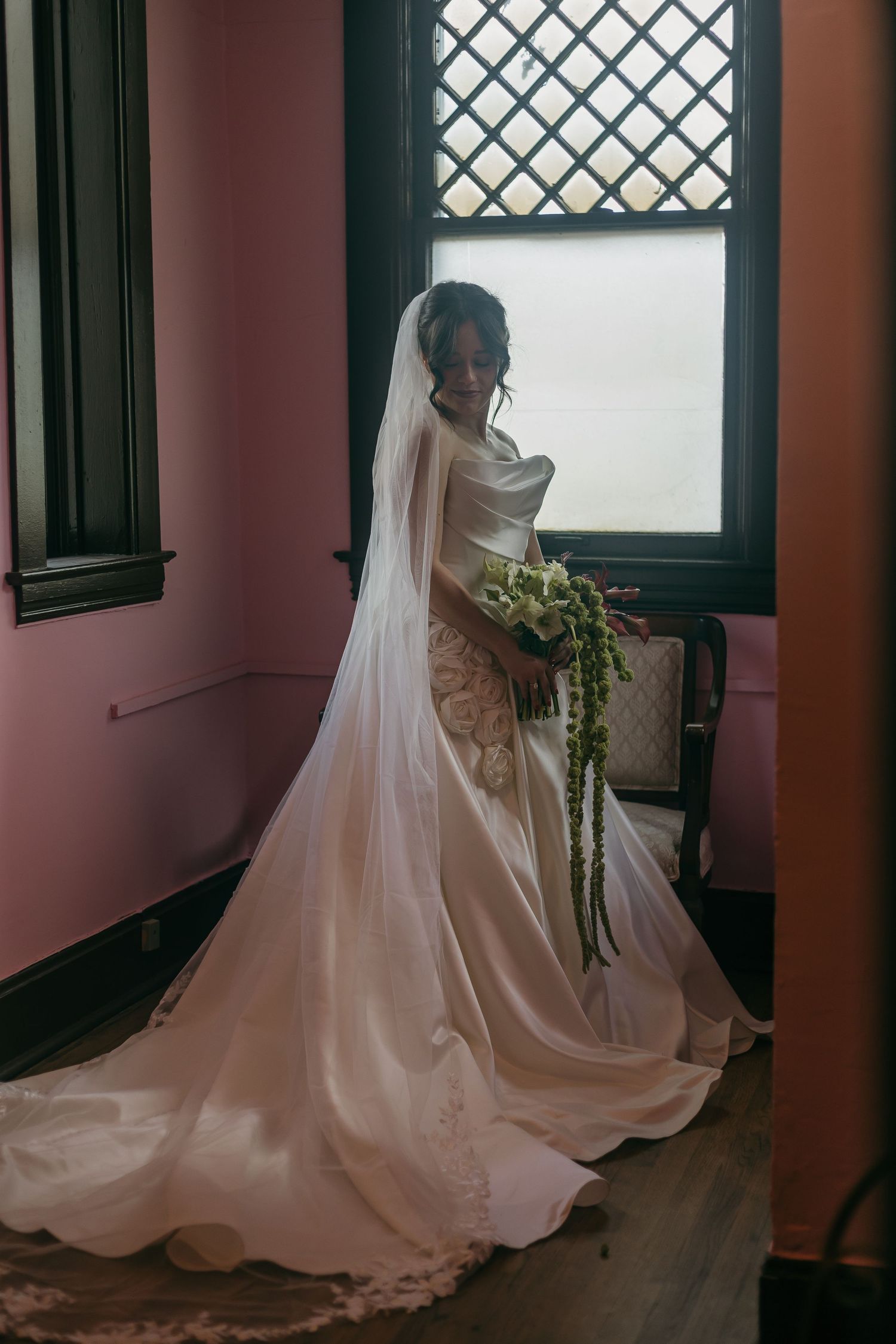 Wedding dress with flowing train against latticed windows in a pink-walled room.