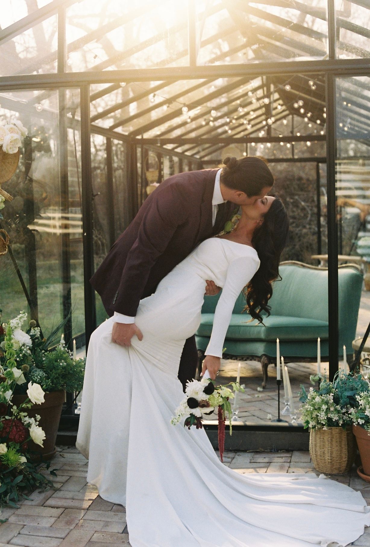 Wedding couple arranging a long white dress train in a sunlit greenhouse setting.