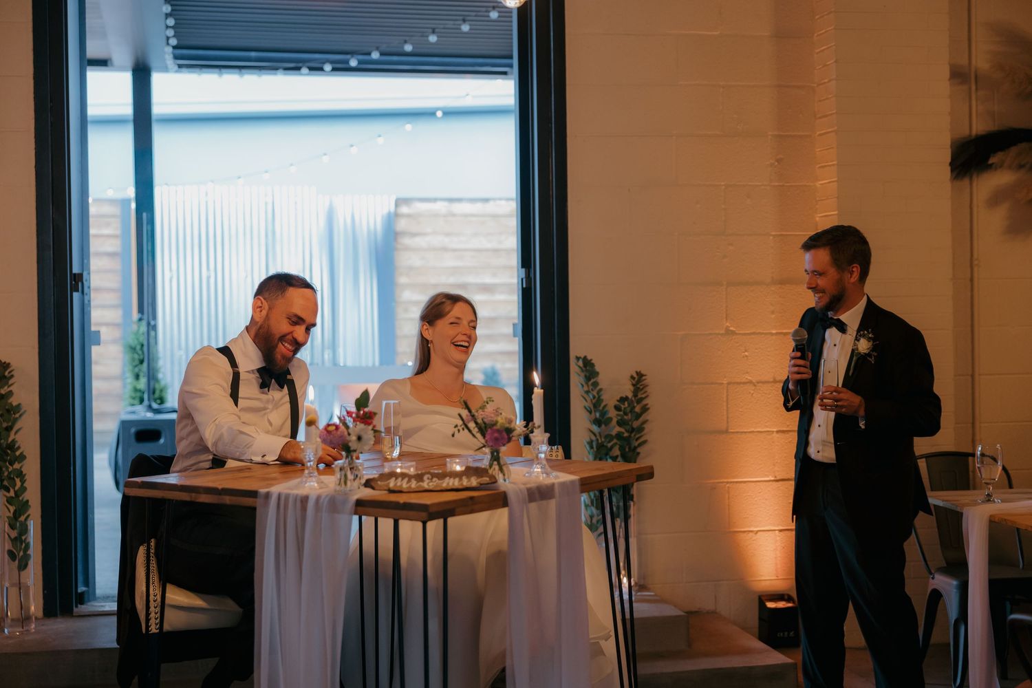 Bride and groom laughing at sweetheart table while groomsman delivers speech at intimate wedding reception.
