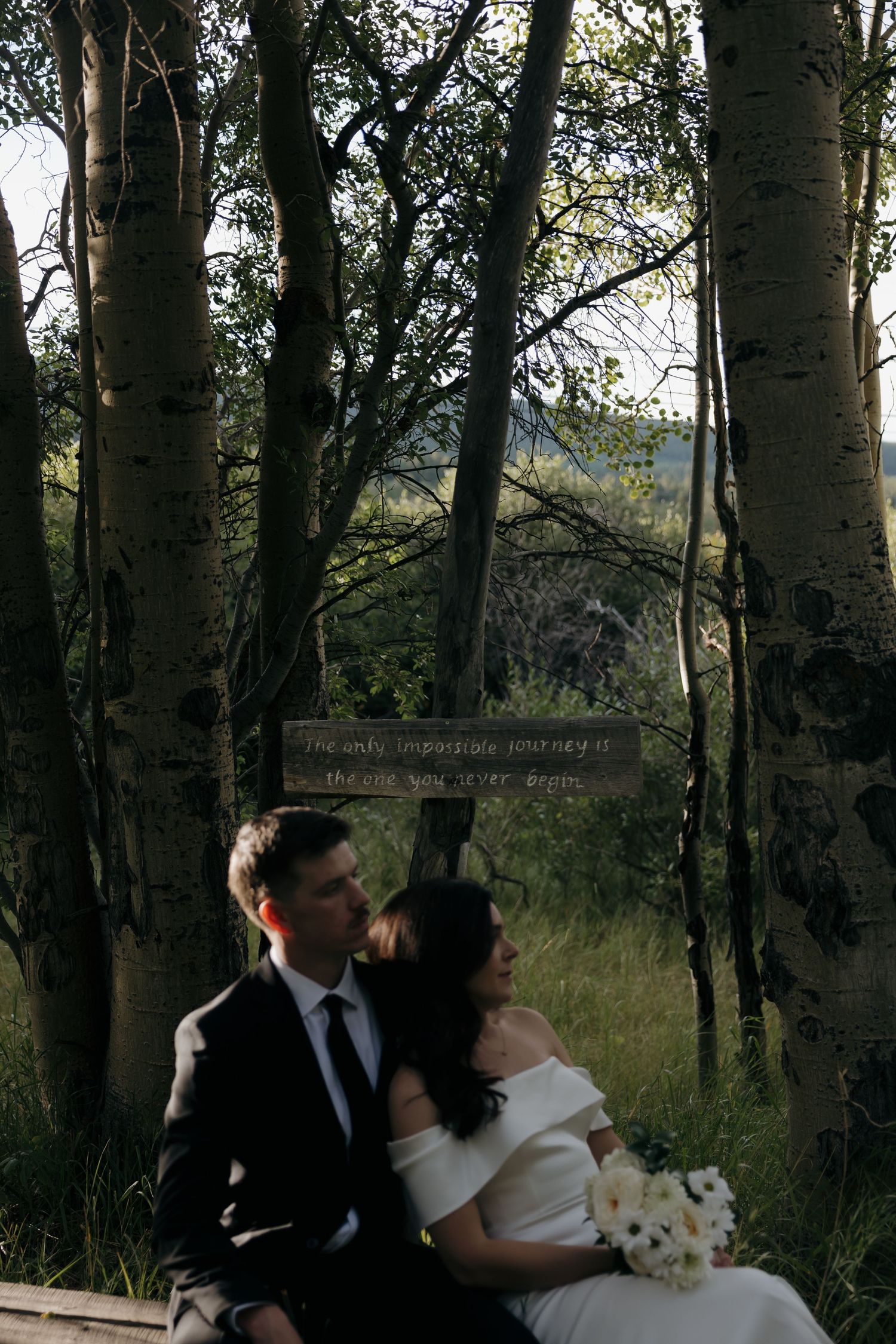 Bride and groom sitting together among aspen trees with inspirational wooden sign at outdoor wedding.