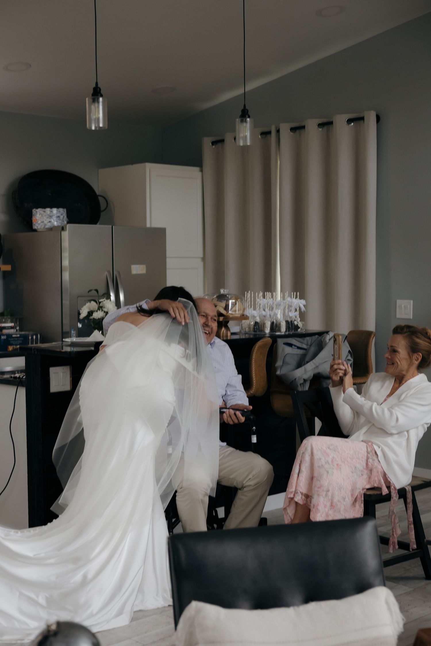 A bride in a white wedding gown and veil embraces a seated man while a woman in a robe photographs the moment indoors.
