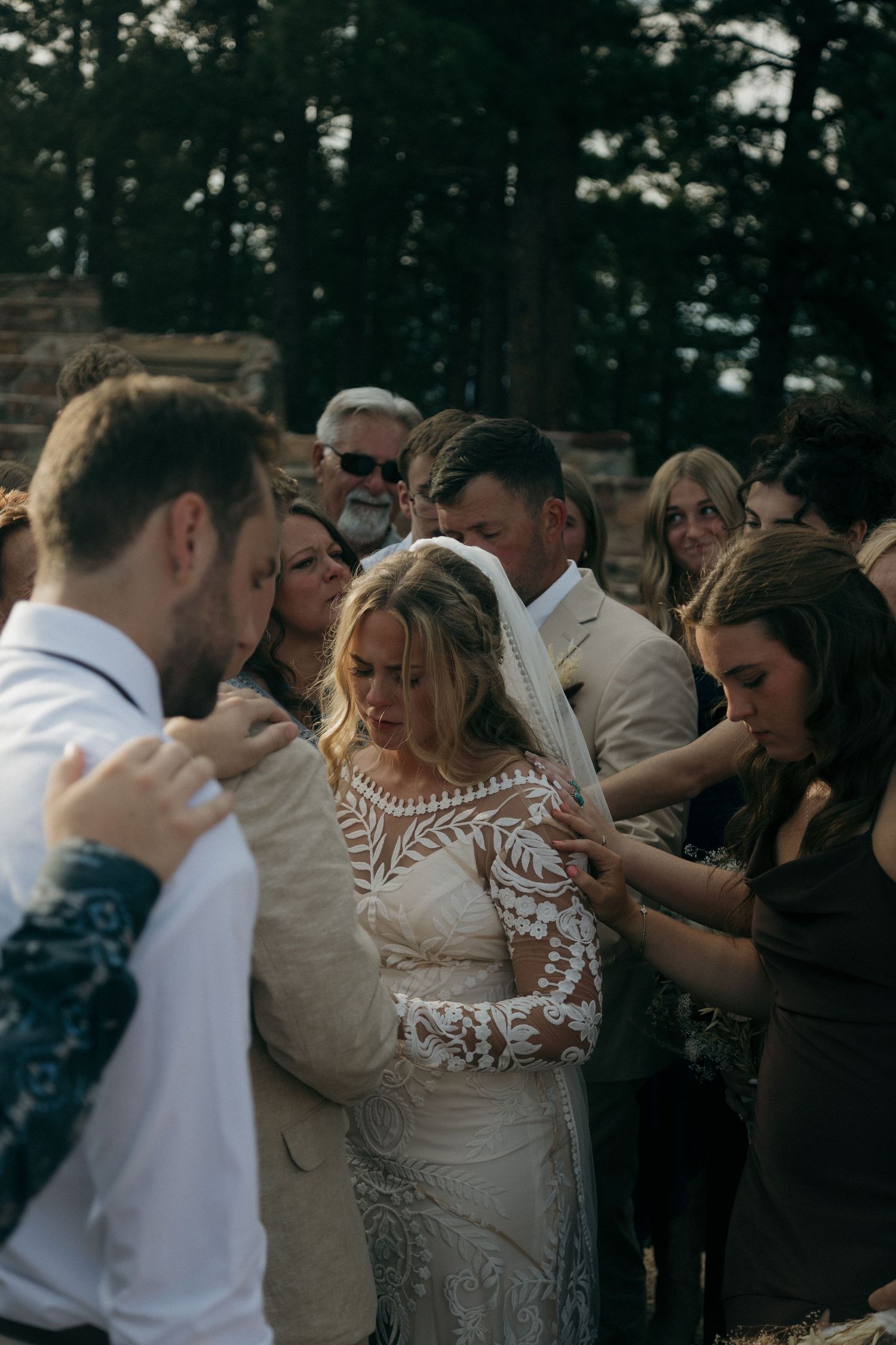 Wedding guests gather around bride and groom in lace gown for outdoor prayer ceremony blessing.