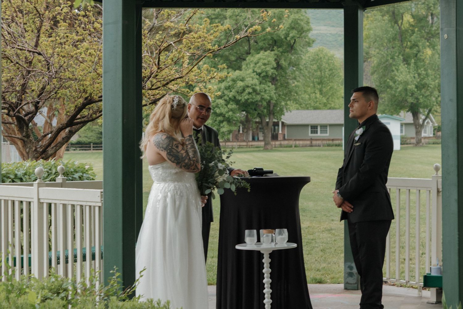 A wedding ceremony takes place under a green gazebo with a black ceremonial table.