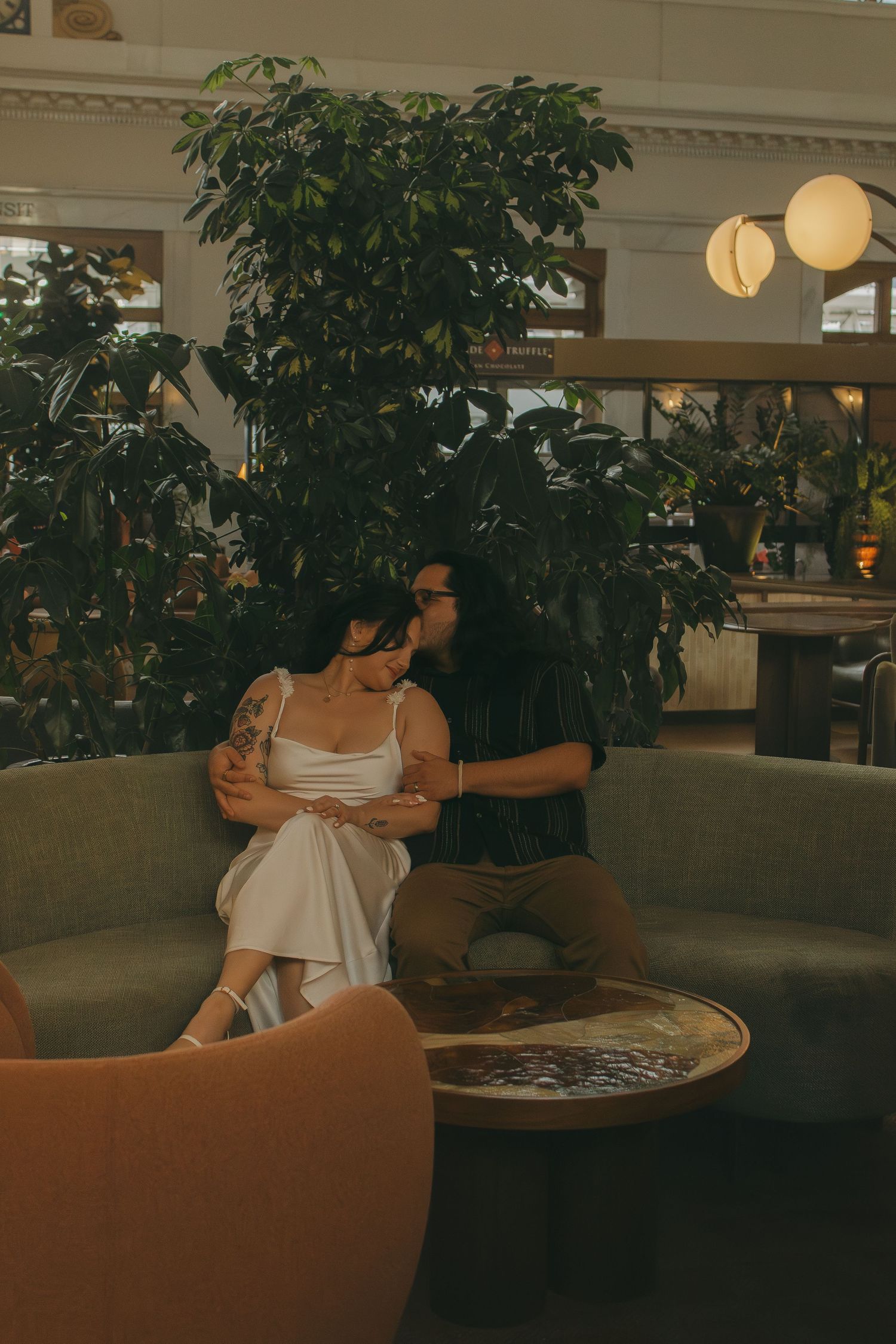 Couple sitting together on curved green sofa with indoor plants and warm lighting.