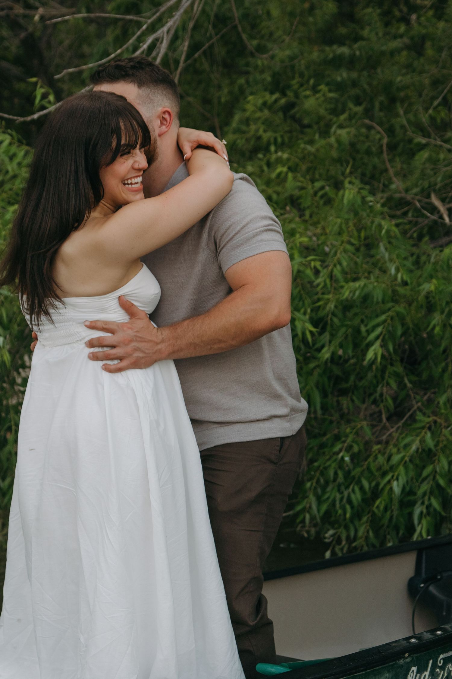 Couple embraces outdoors in casual attire against a natural green backdrop.