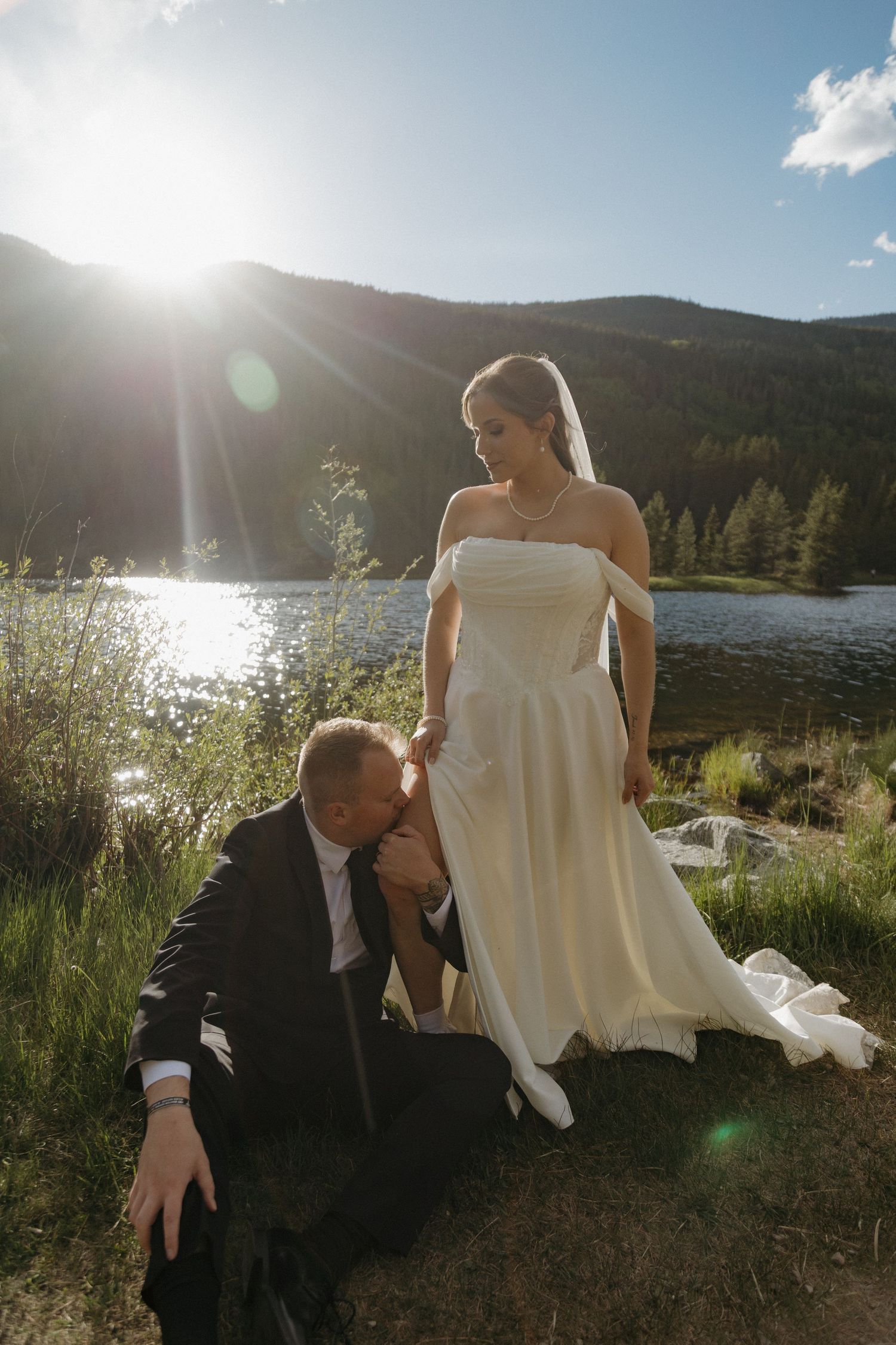 Groom kneeling to kiss bride's hand by mountain lake with bright sun flare overhead.