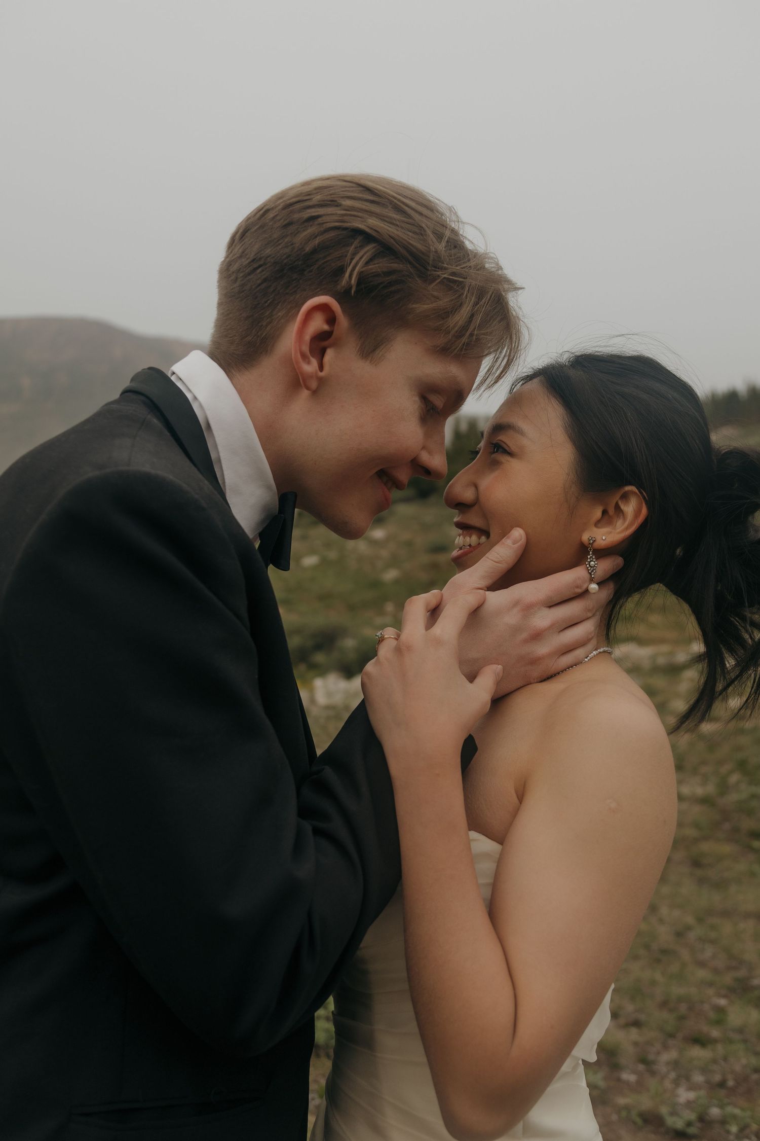 Bride and groom share an intimate moment touching foreheads and smiling on a misty mountainside.