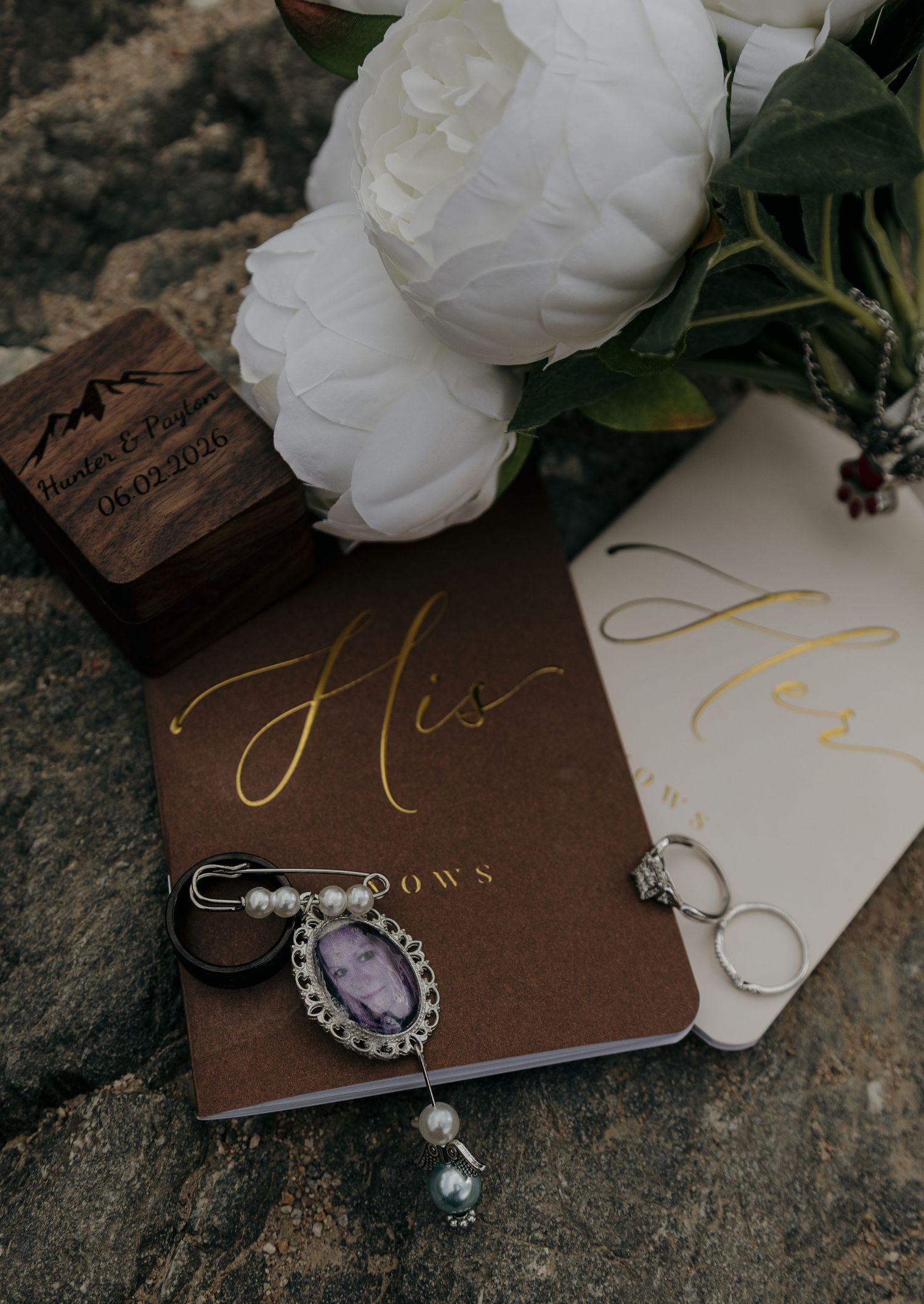 Wedding flat lay with His and Hers vow books, rings, bouquet, and memorial charm on rustic stone.