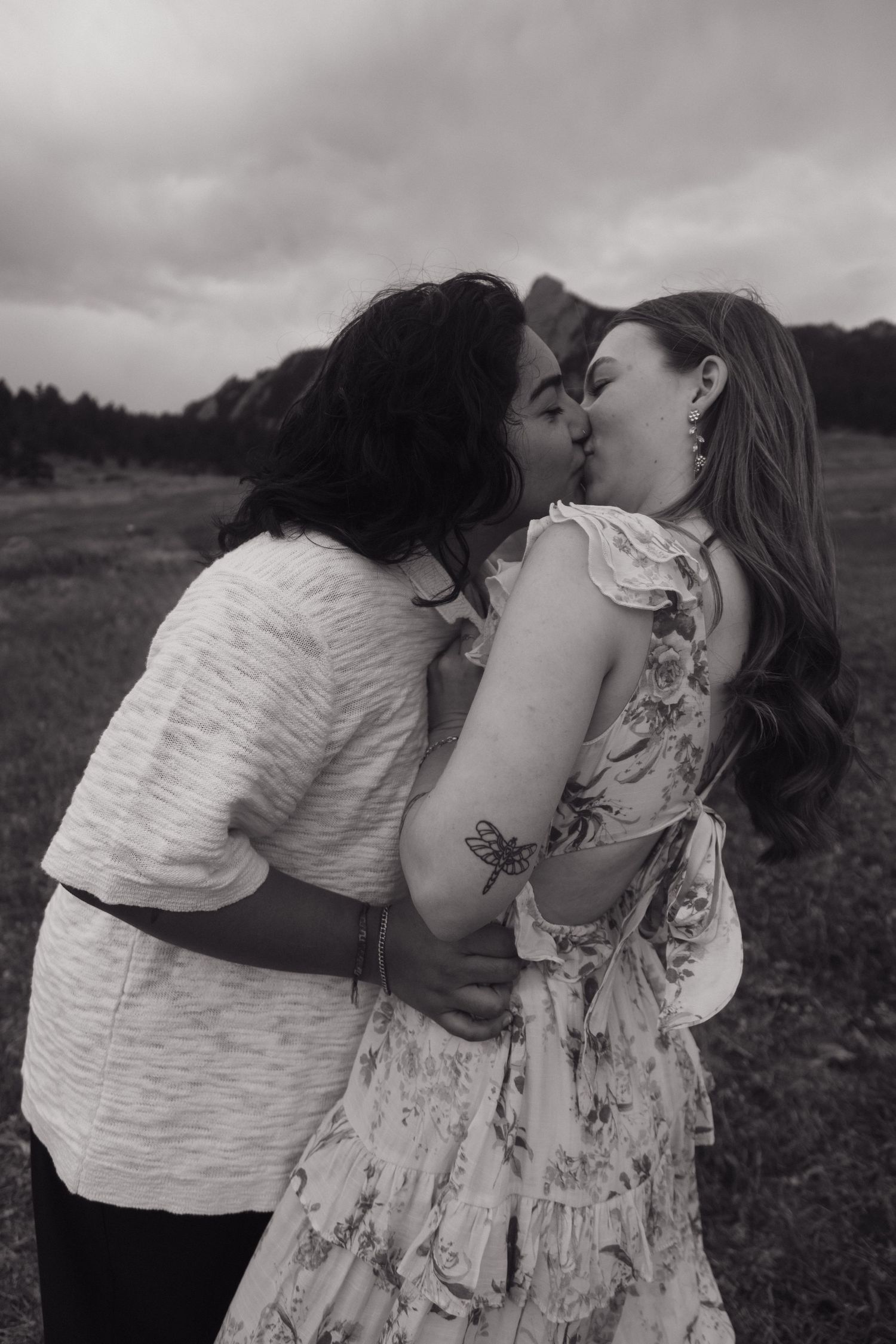 Black and white photo of couple sharing a kiss in a field with dramatic cloudy sky backdrop.