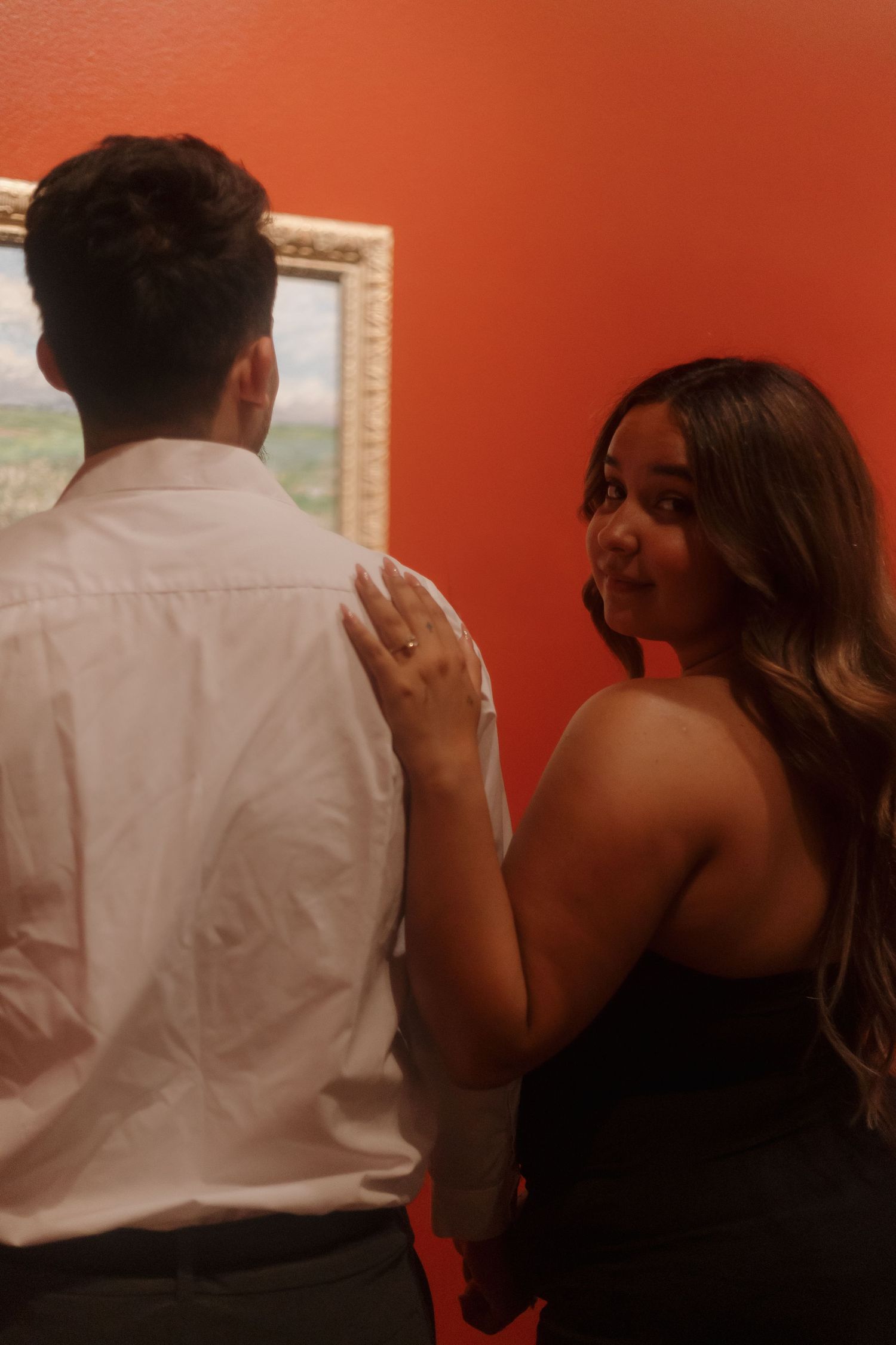 Couple holding hands in front of a framed painting on an orange wall, woman smiling over her shoulder.