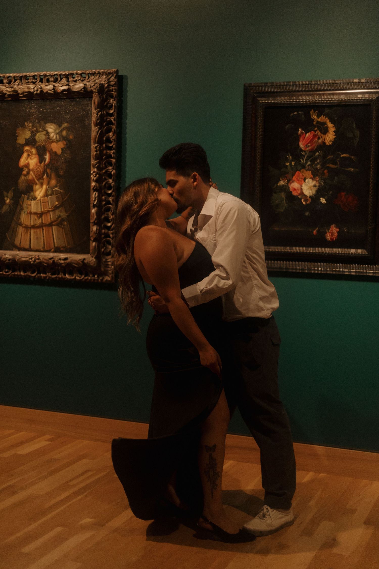 Couple sharing a passionate kiss in an art museum gallery surrounded by ornate floral still-life paintings.