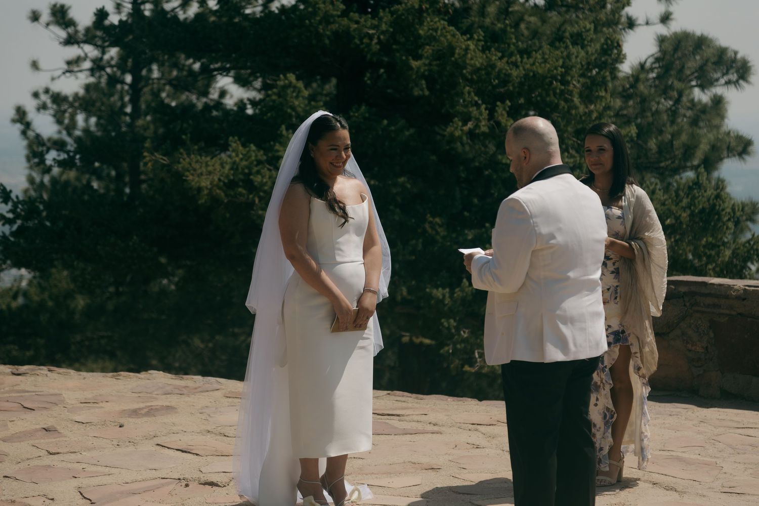Bride in white dress and veil exchanges vows with groom at outdoor wedding ceremony surrounded by trees.