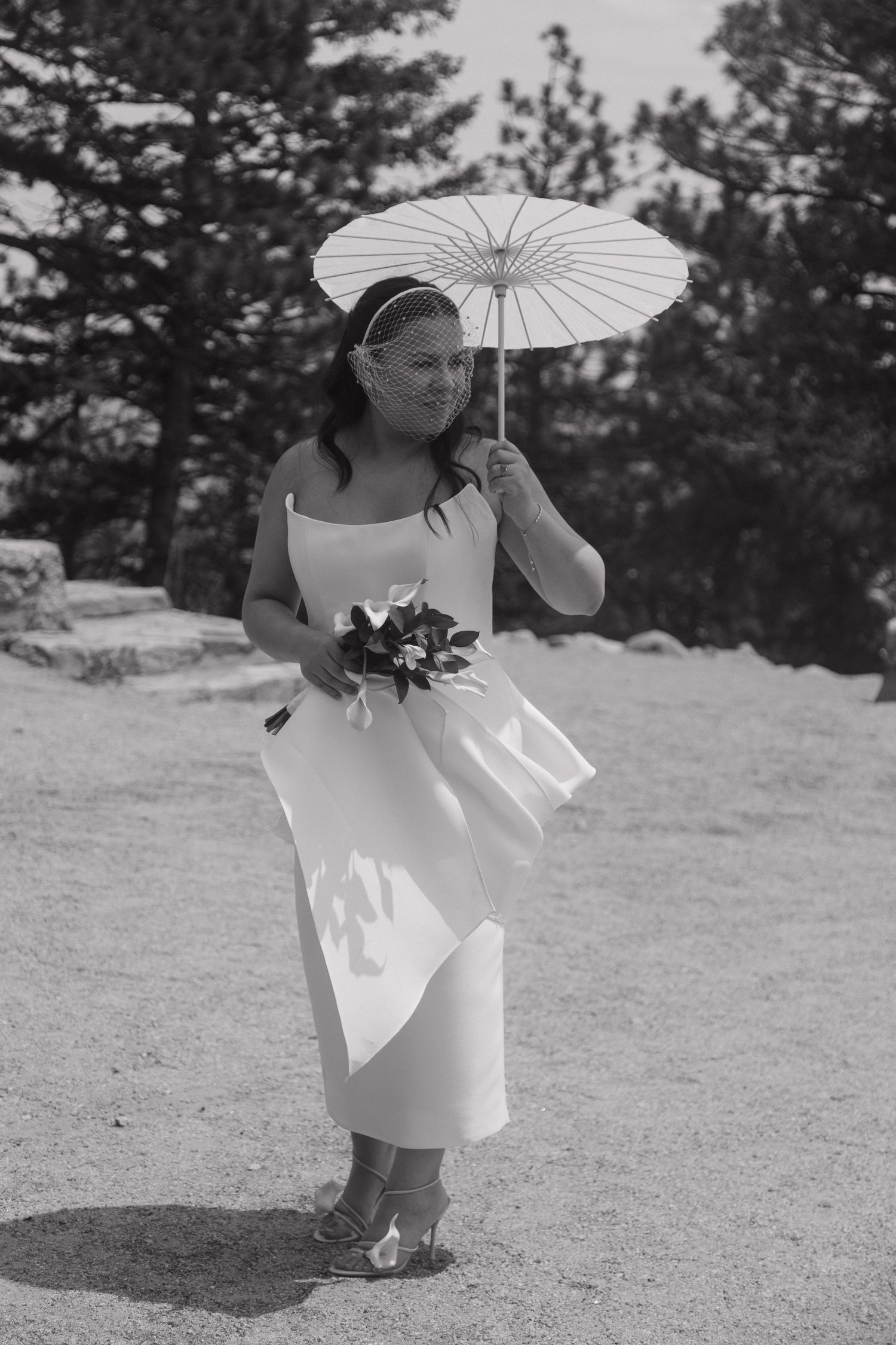 Bride in white dress holds parasol and bouquet at outdoor mountain wedding ceremony, black and white.