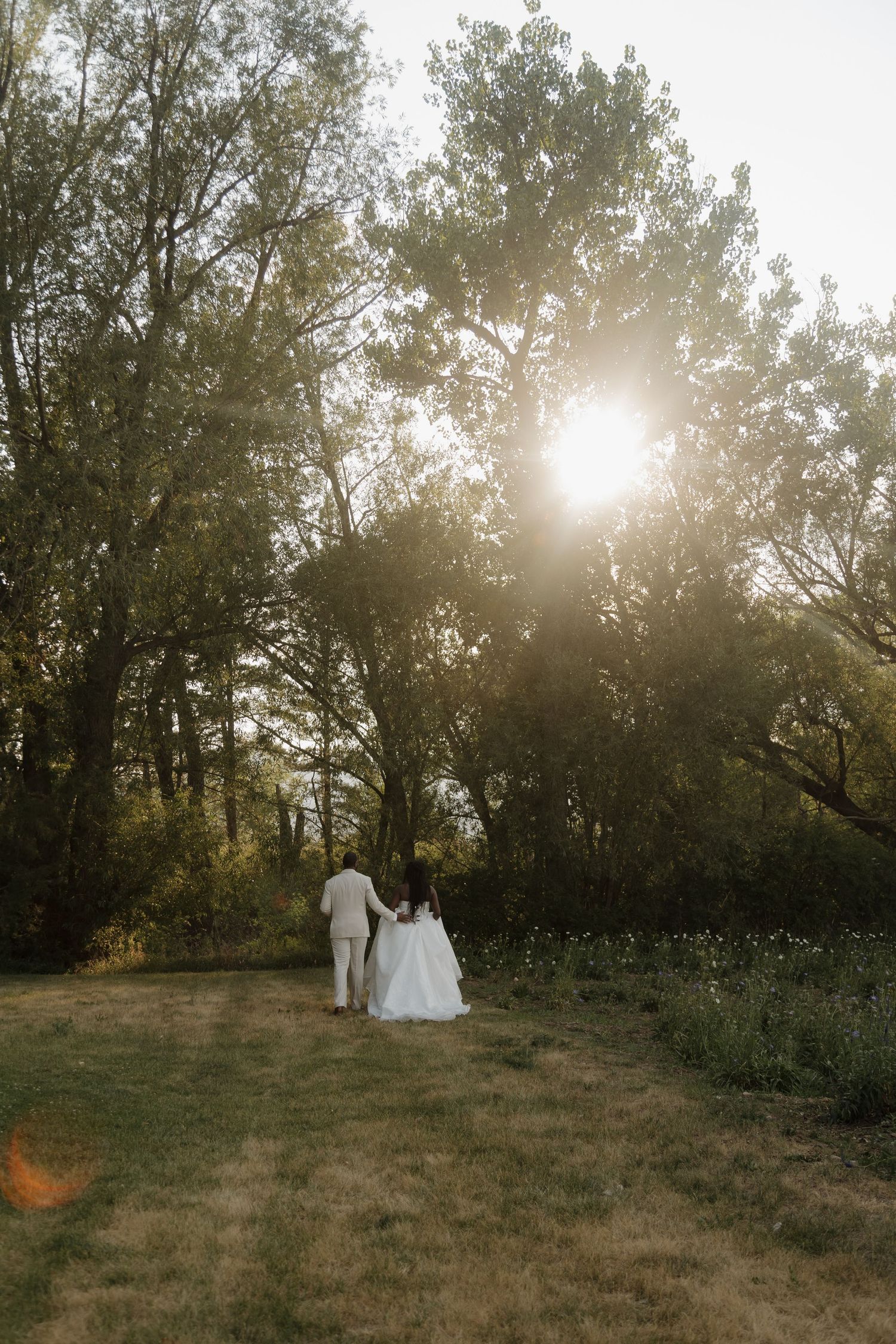 Bride and groom walking hand in hand toward sunlit trees in a serene garden at sunset.