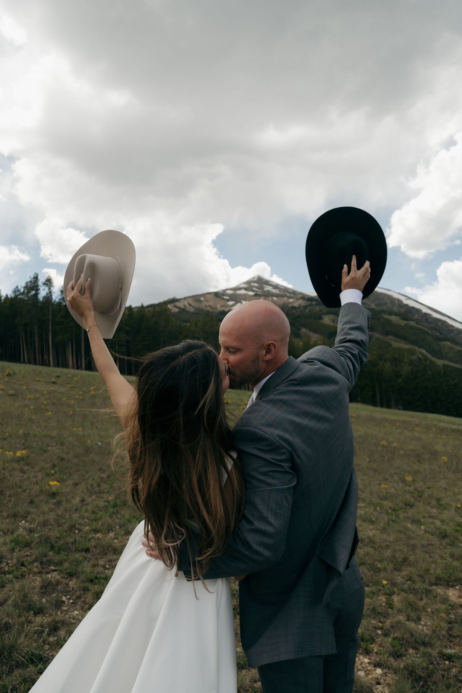 A bride and groom kiss while raising cowboy hats in a scenic mountain meadow.
