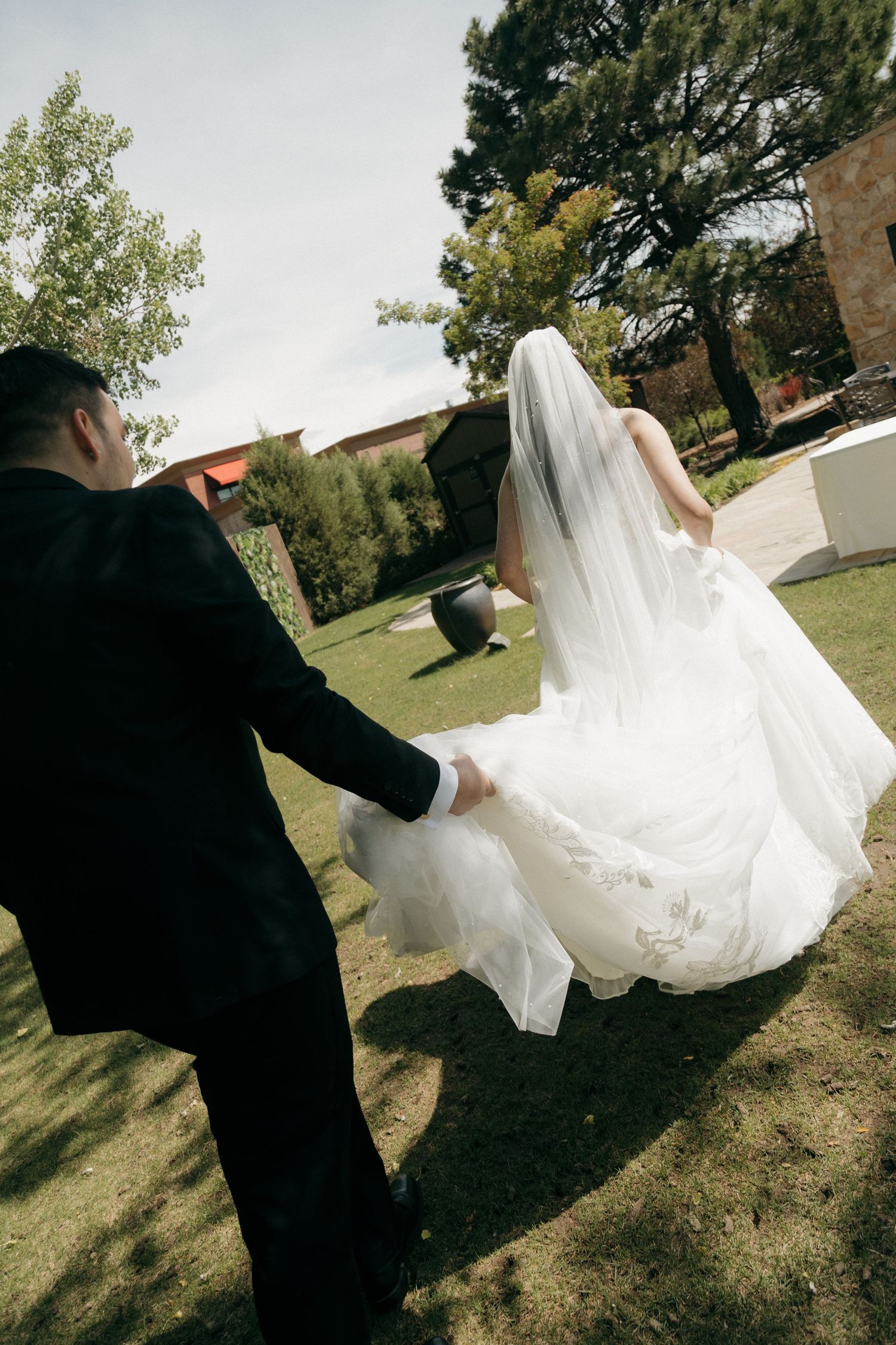 Bride in white gown and veil walks hand-in-hand with groom in black suit through lush garden on wedding day.