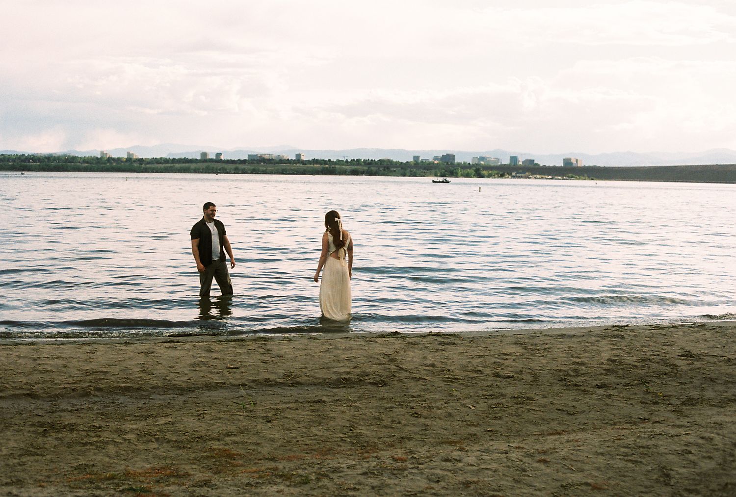 A couple stands at the water's edge on a sandy beach, the bride in a flowing white gown.