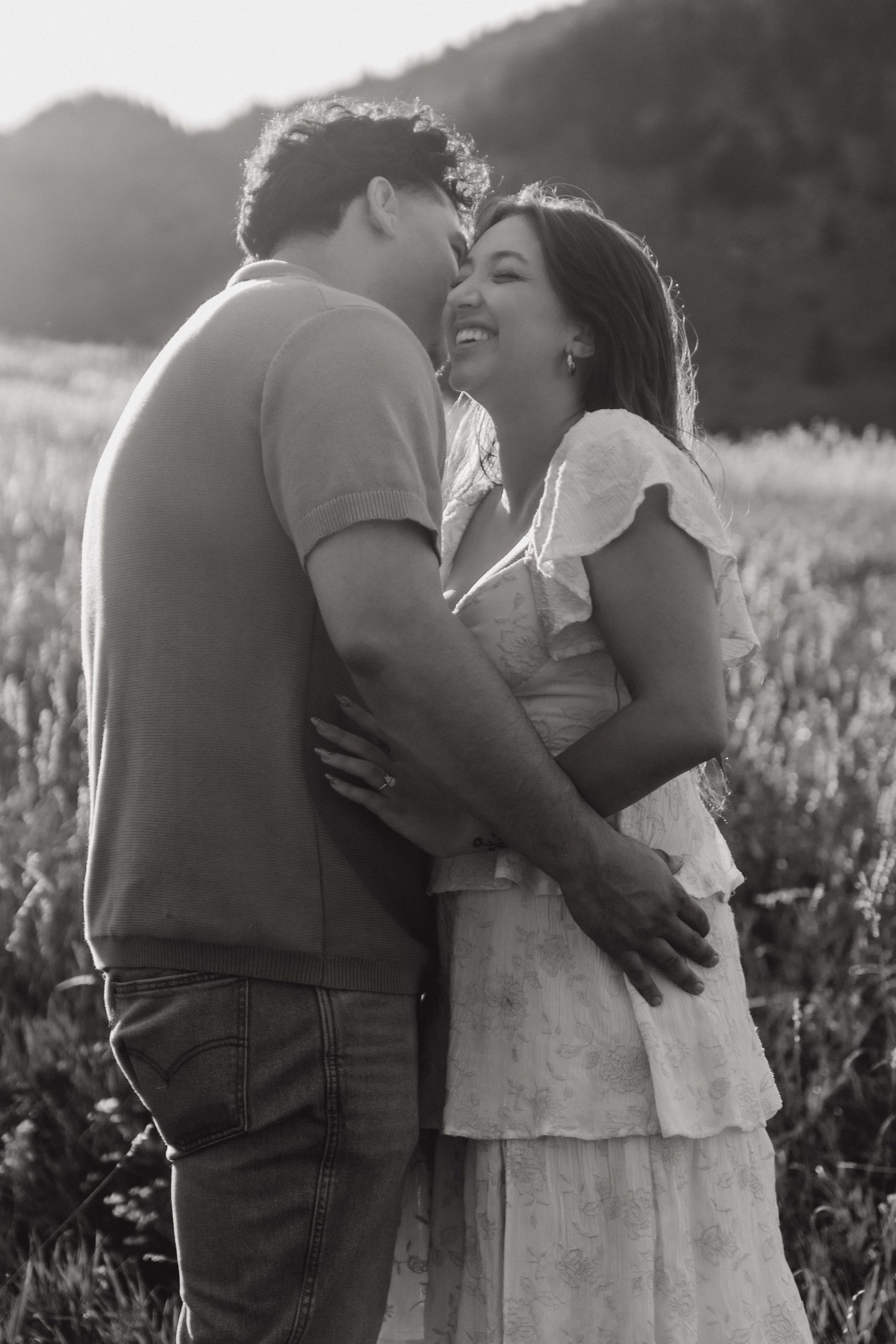 Black and white photo of laughing couple embracing in sunlit meadow during engagement session.