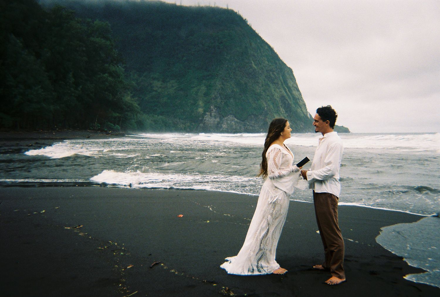 Couple exchanging vows on a black sand beach with dramatic ocean waves and lush green cliffs in Hawaii.