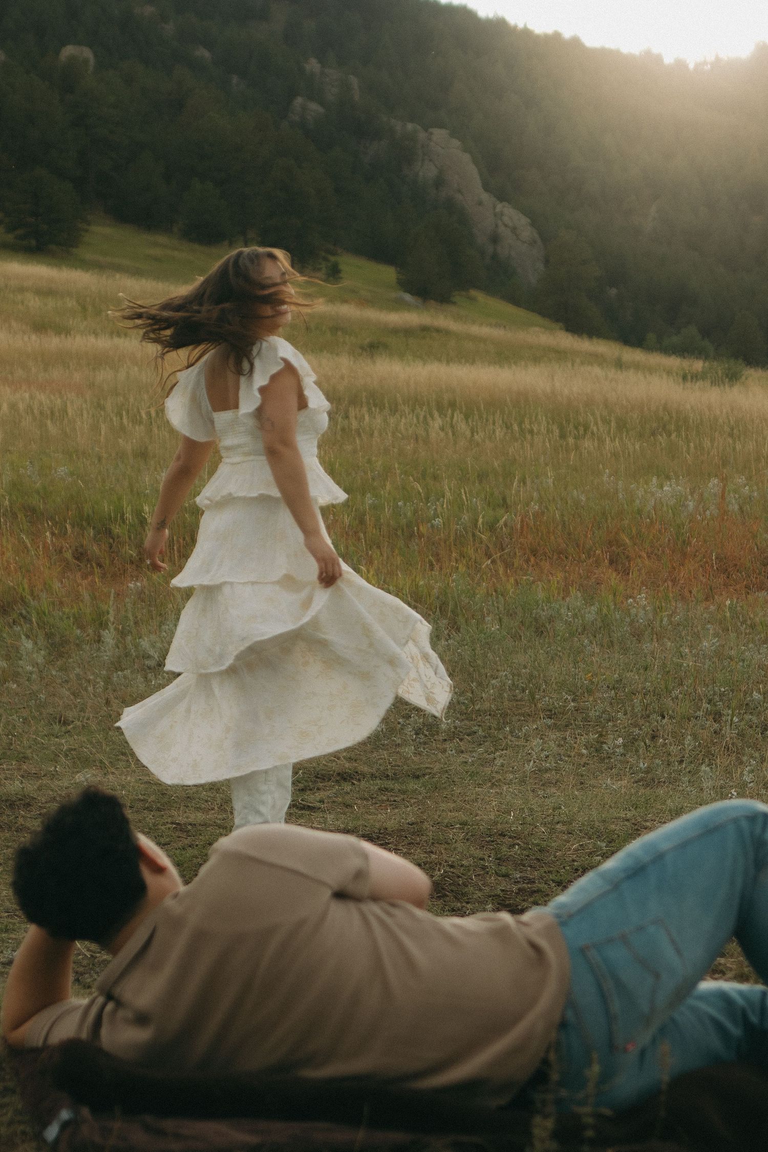 Woman in white tiered dress twirling in sunlit meadow while man relaxes on grass below.
