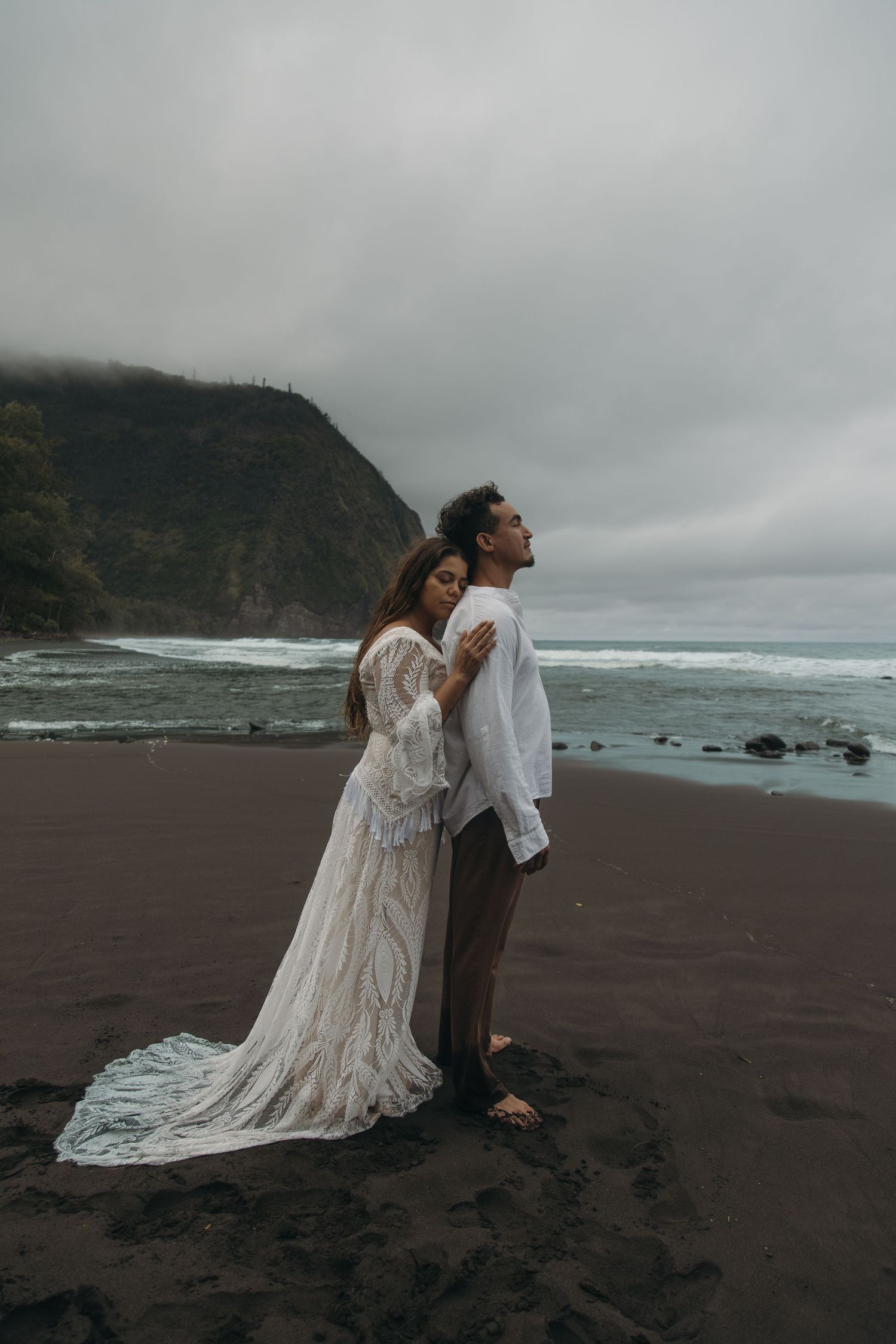 Bride in lace gown embracing groom from behind on dramatic black sand beach with ocean cliffs.