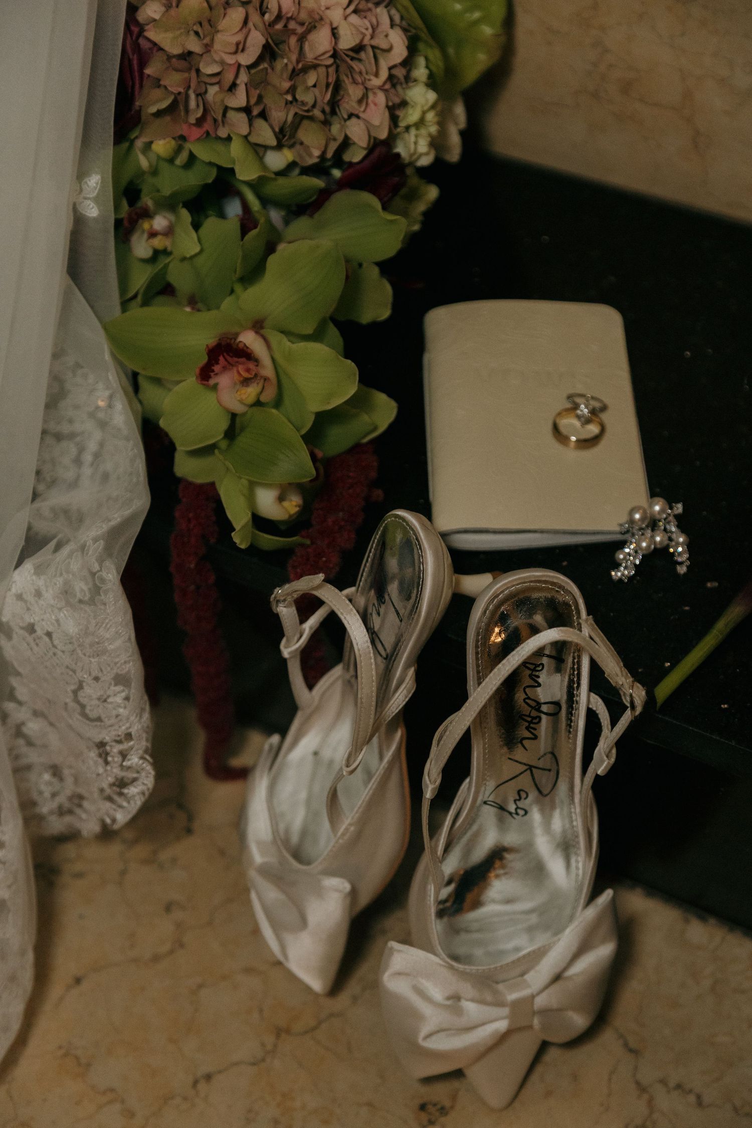 White satin bridal heels with bow detail beside wedding rings, bouquet of hydrangeas and orchids on marble surface.
