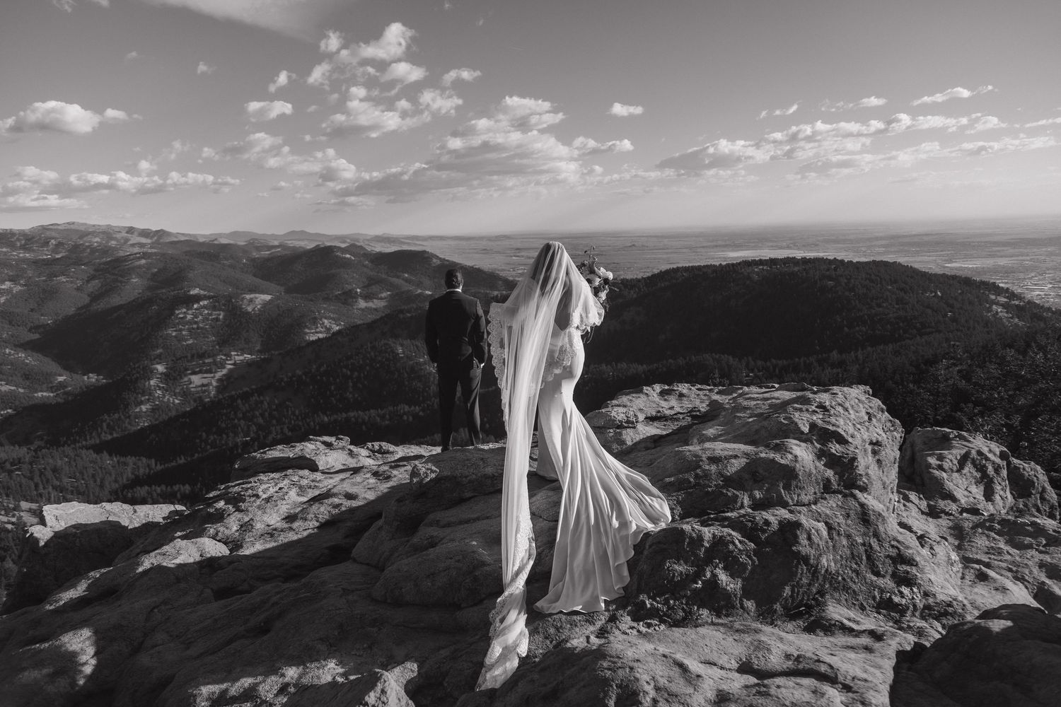 Black and white photo of bride in flowing veil standing on mountain summit with groom.