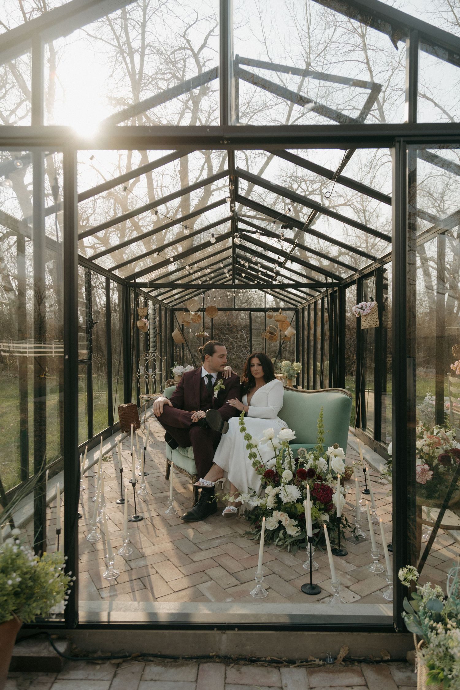 Bride and groom seated together inside a sunlit glass greenhouse surrounded by floral arrangements.