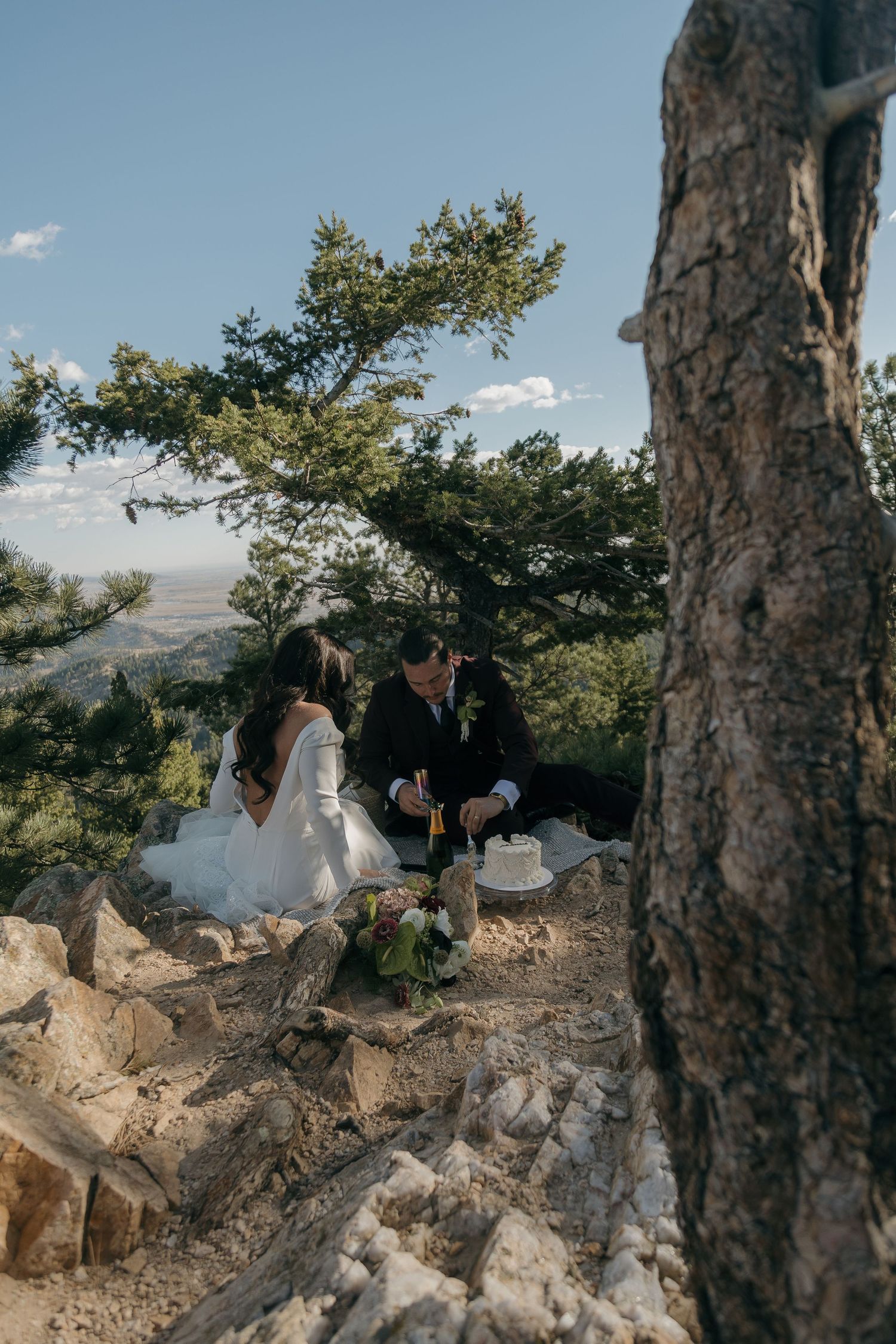 Bride and groom sitting on rocky mountain summit sharing champagne and wedding cake outdoors.