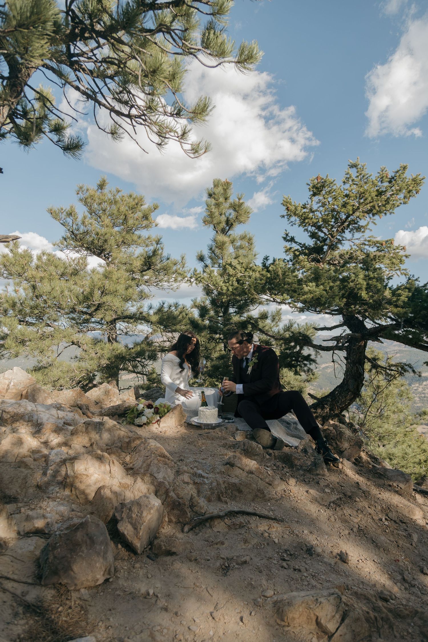 Couple sitting among pine trees on rocky mountain terrain during outdoor wedding elopement celebration.