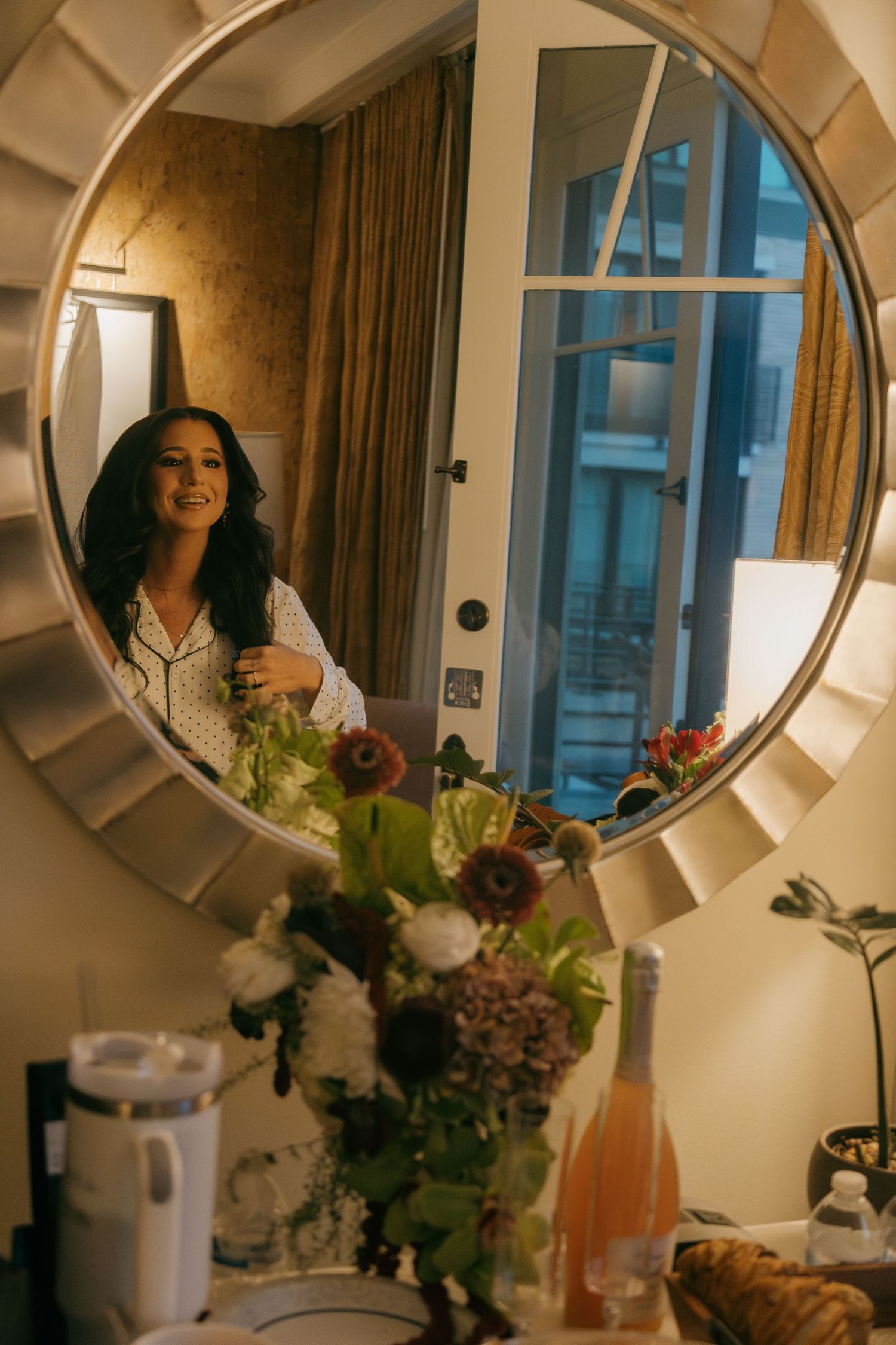 Bride in white robe smiles in round mirror reflection surrounded by wedding flowers and champagne.