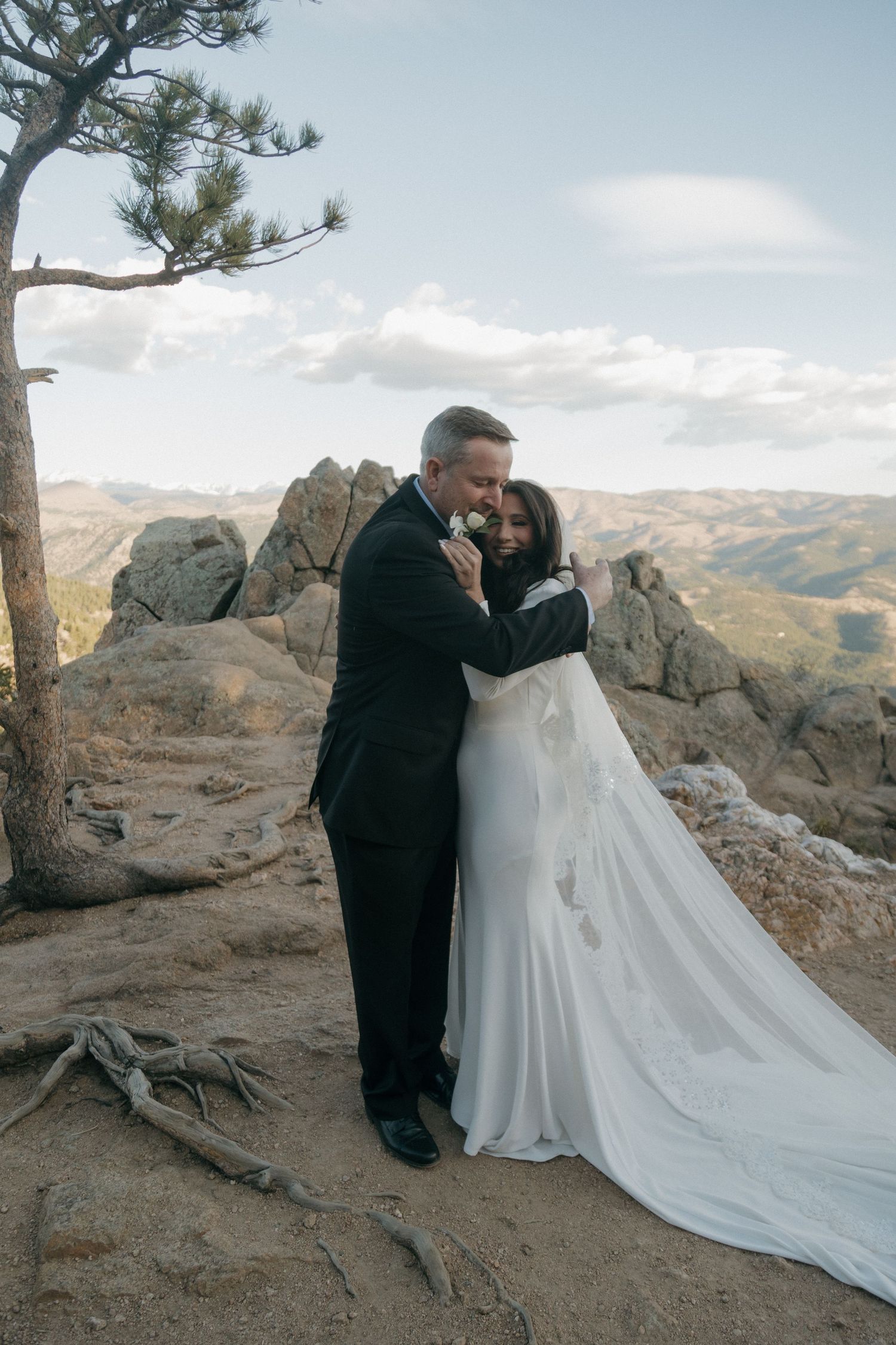 Bride and groom embrace on a rocky mountain overlook, she in a flowing white gown with veil, he in a black tuxedo.