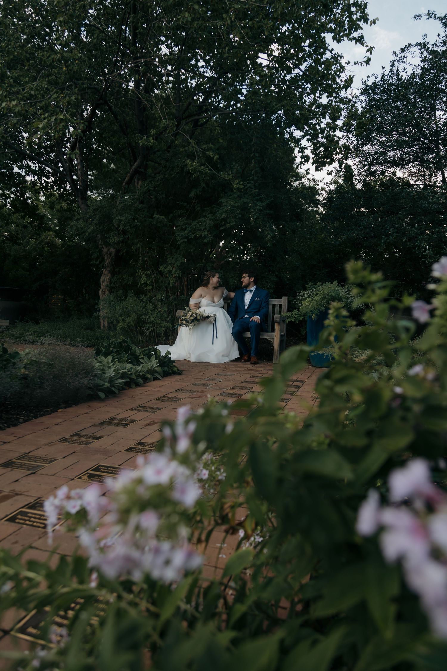 Bride and groom sharing a romantic moment on a brick garden path surrounded by lush green trees at dusk.