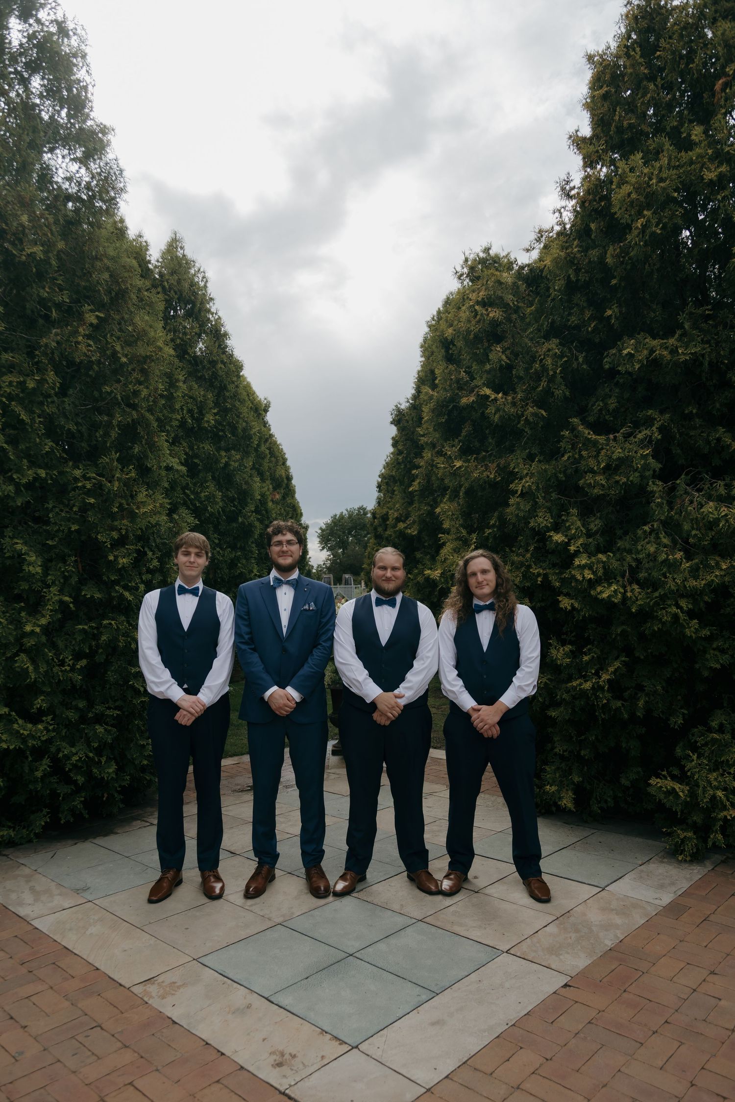 Four groomsmen in navy blue suits and bow ties posing formally between tall green hedges at a wedding.
