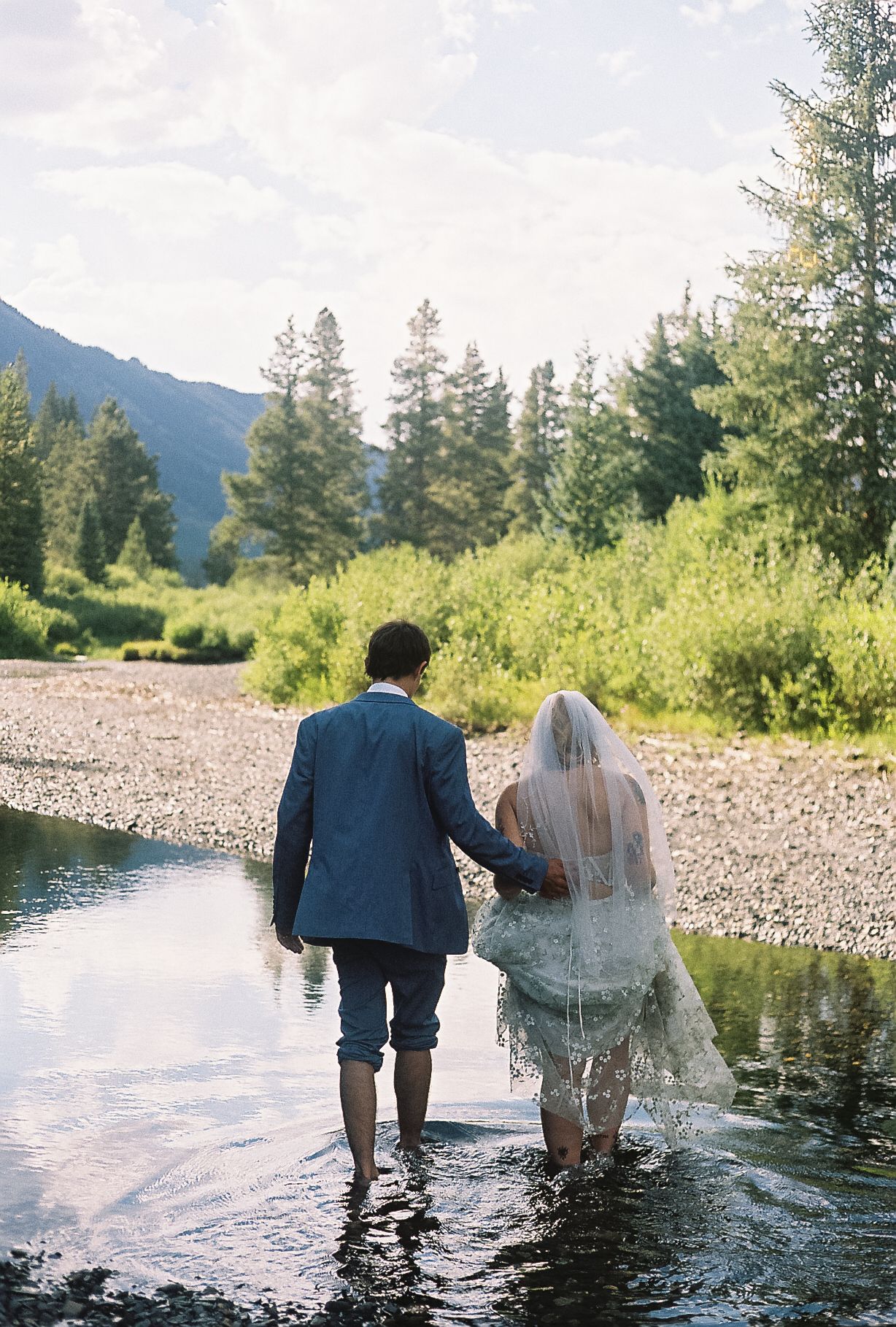 Bride and groom wade hand in hand through a shallow mountain stream surrounded by lush green trees and hills.