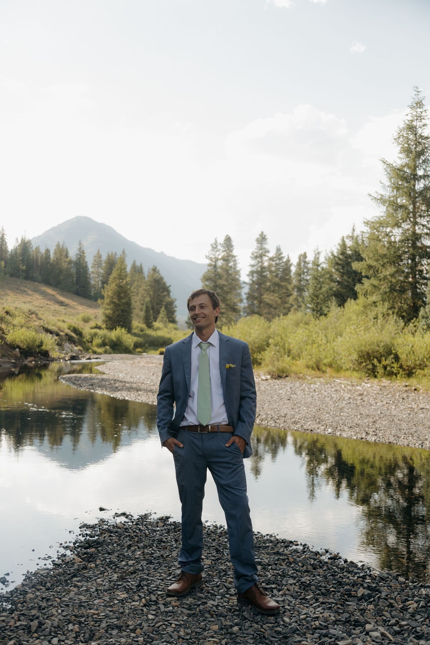 Groom in blue suit standing confidently by a reflective mountain stream with pine trees backdrop.