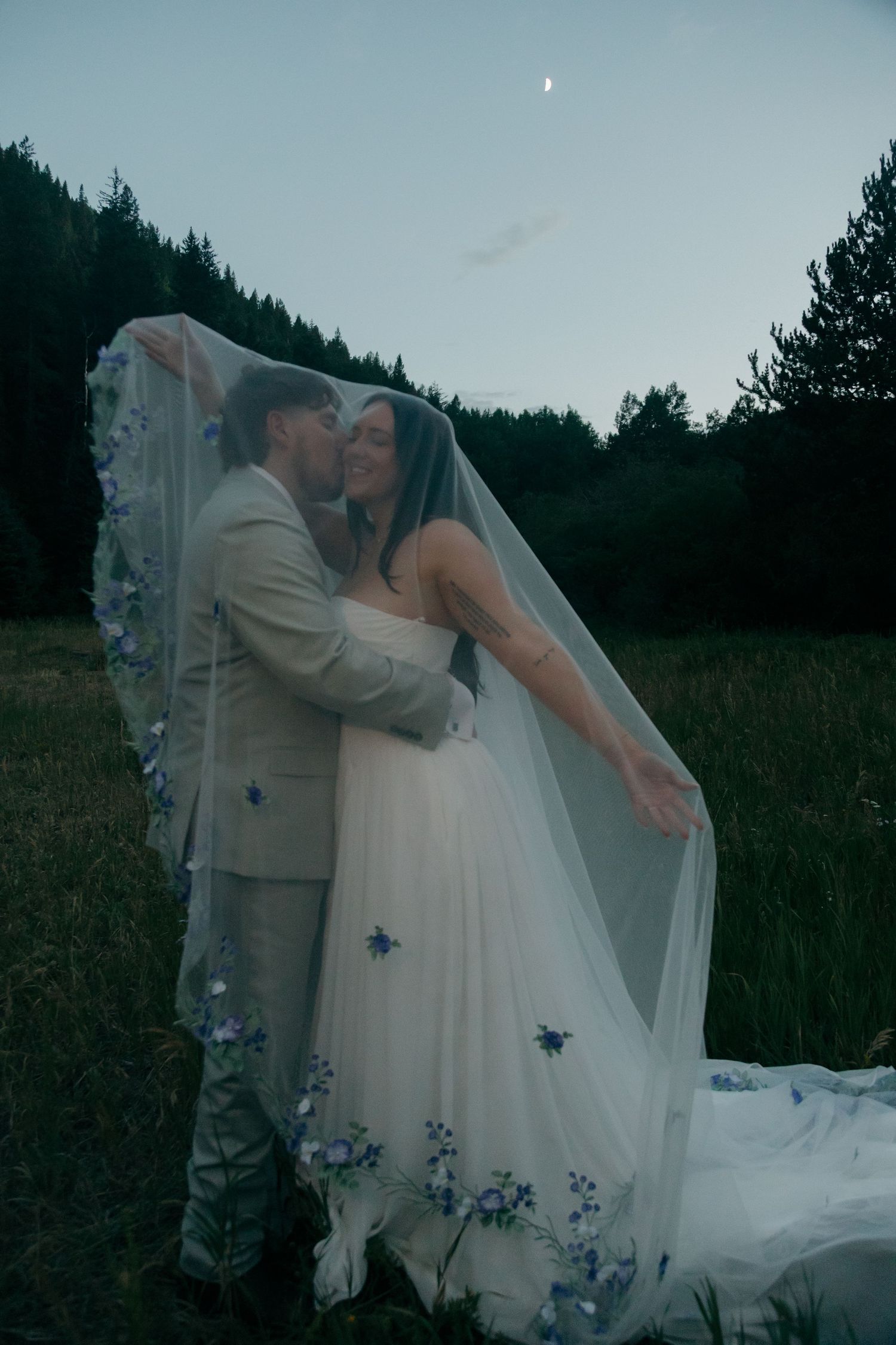 Bride and groom embracing under floral veil in meadow at twilight with crescent moon above.