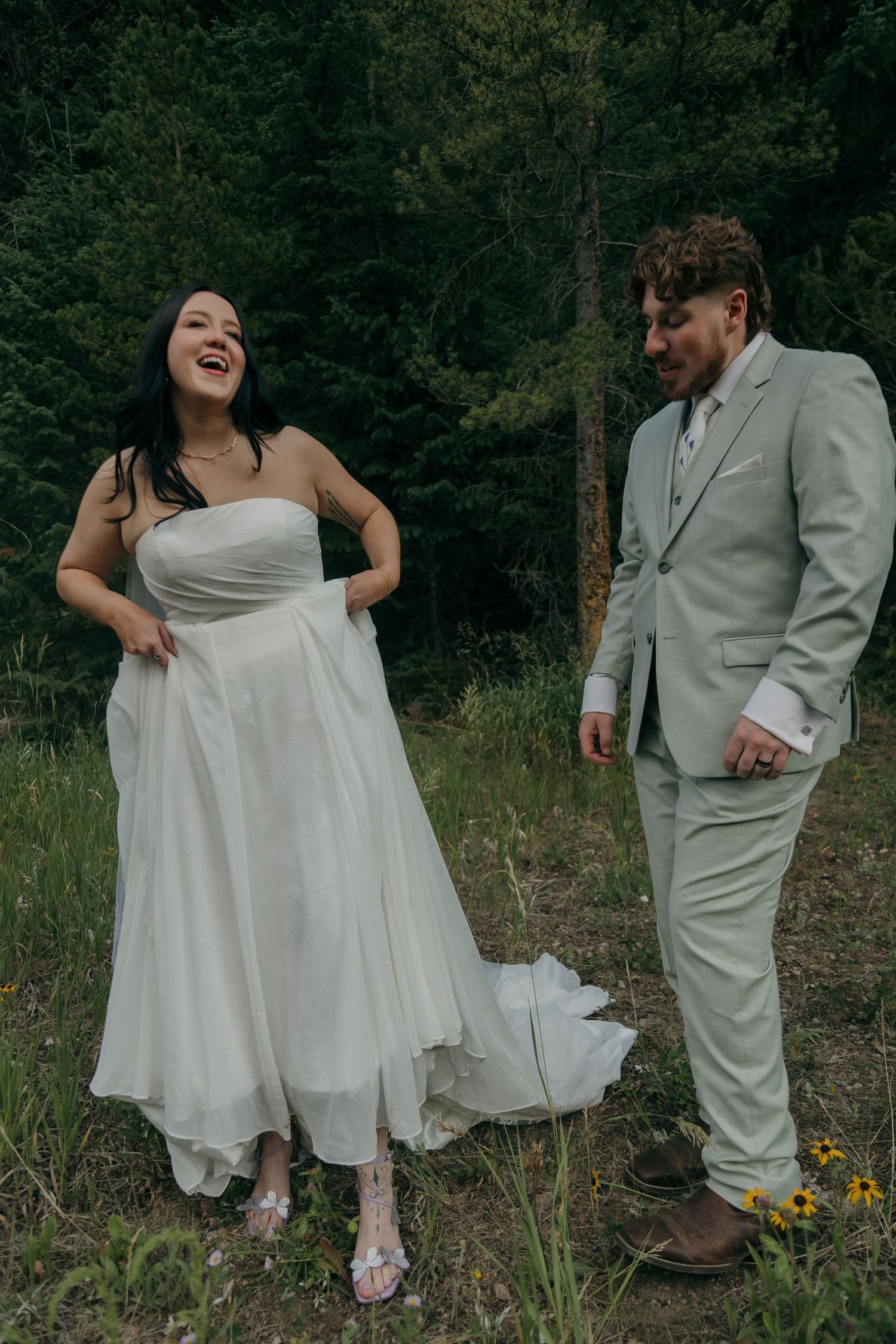 Laughing bride in strapless white gown and groom in sage suit share joyful moment in forest clearing.