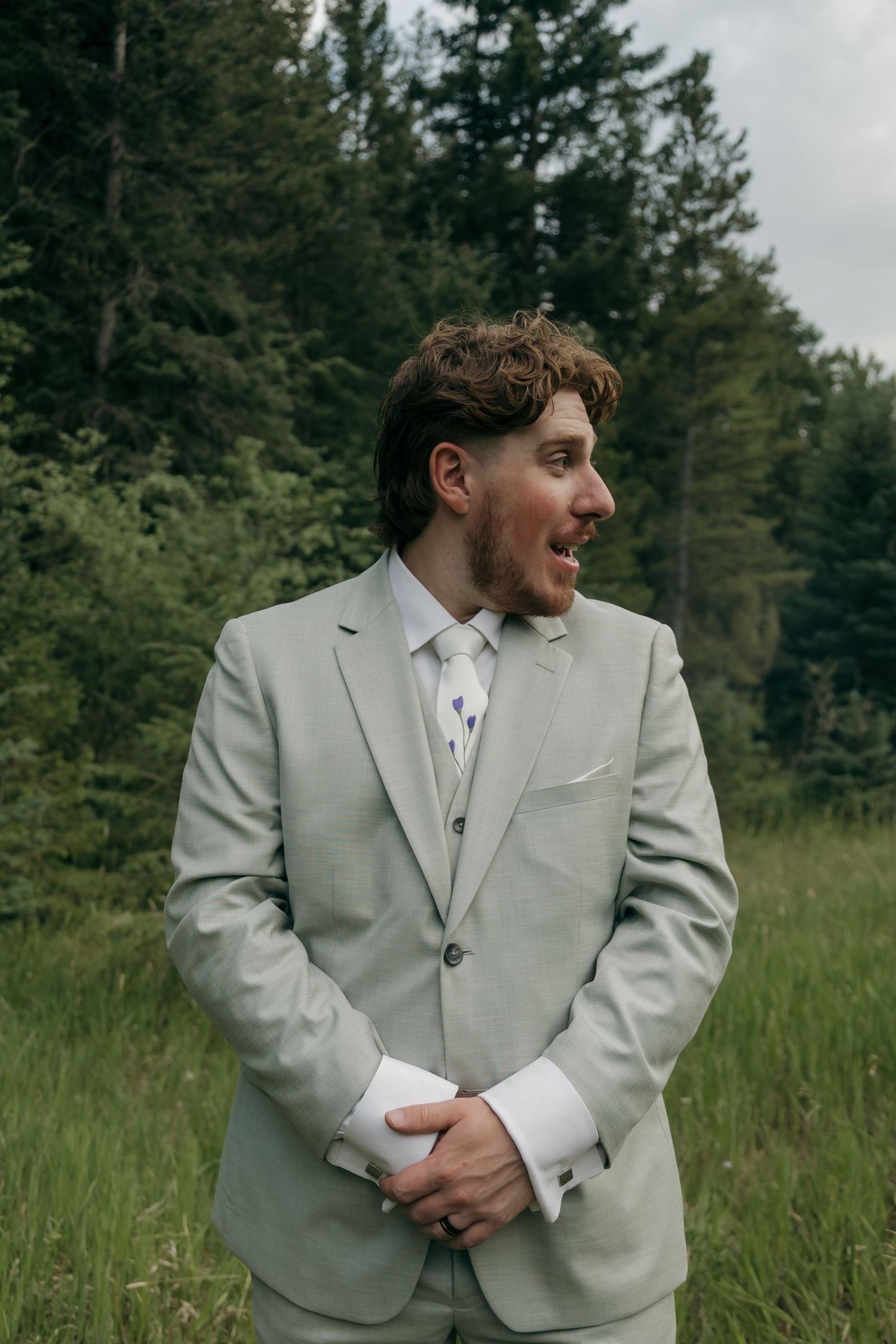 A groom in a light gray suit and tie smiles outdoors surrounded by tall green trees on his wedding day.