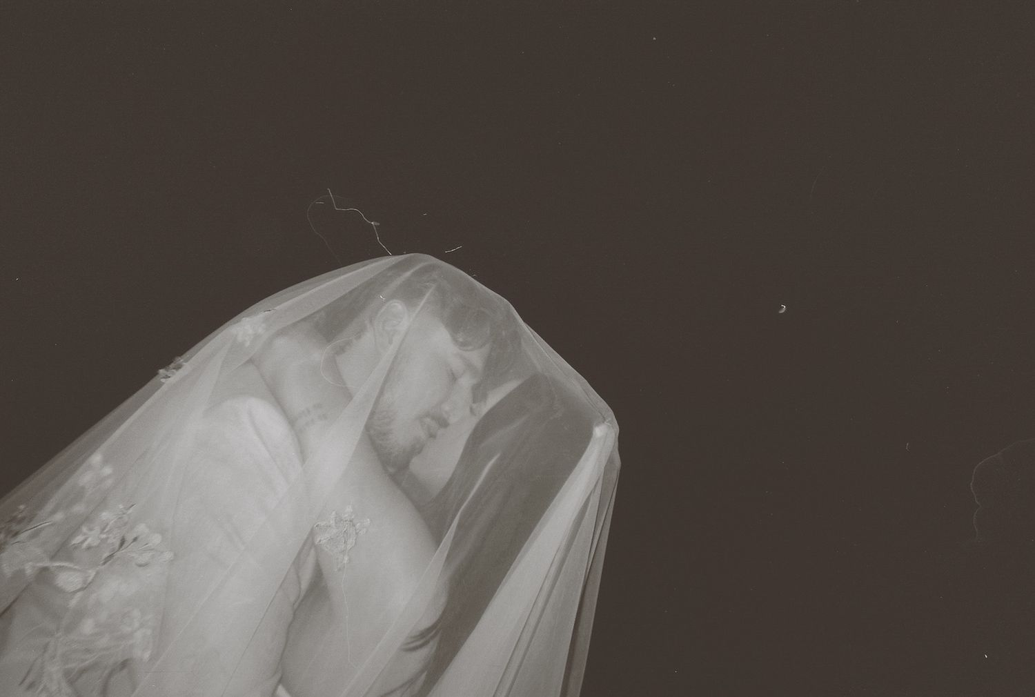 Dreamy overexposed film photo of bride covered in white veil on dark background.