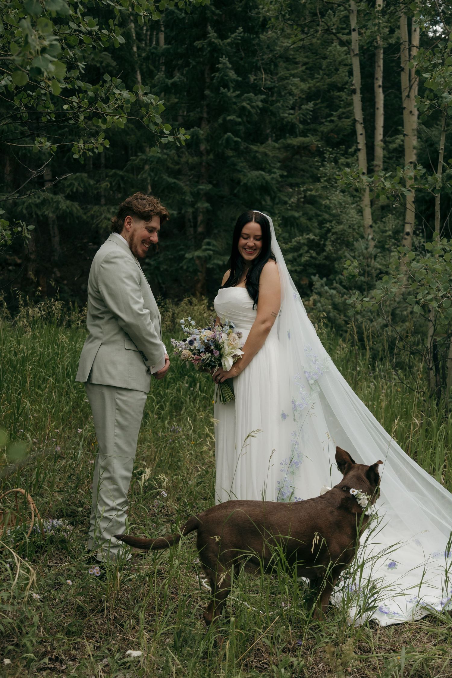 Bride and groom smiling with chocolate dog in lush forest wedding portrait.