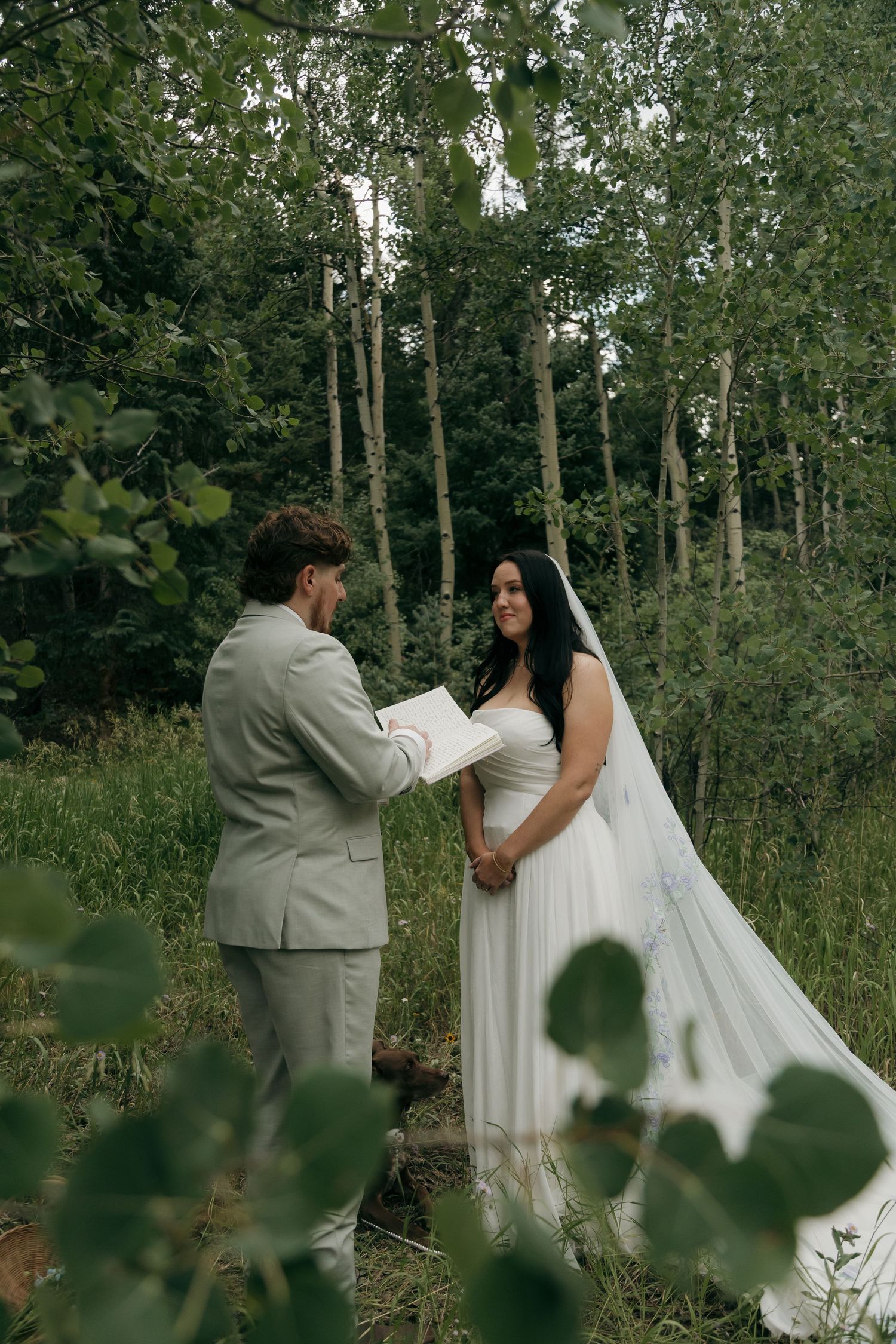 Bride and groom exchange vows in a lush forest setting, groom reading from a book in a gray suit.