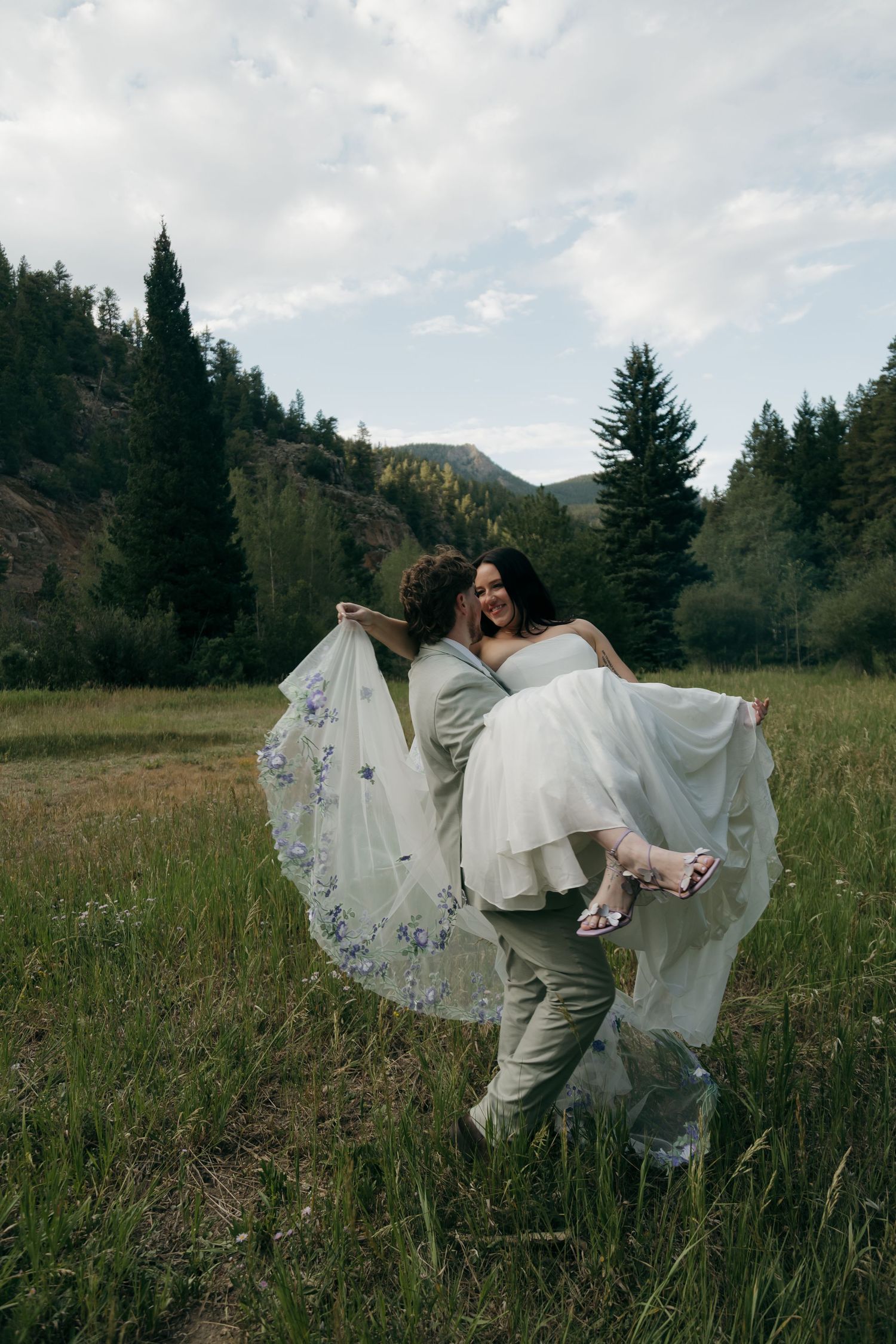 Groom lifts smiling bride in a floral-embroidered wedding gown in a scenic mountain meadow with evergreen trees.
