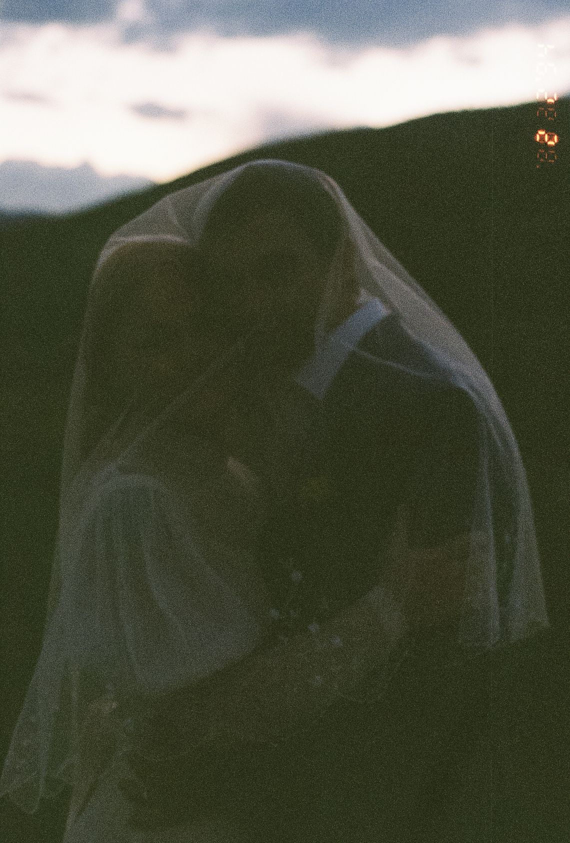Bride and groom share a romantic kiss under a wedding veil at dusk with mountain silhouette backdrop.