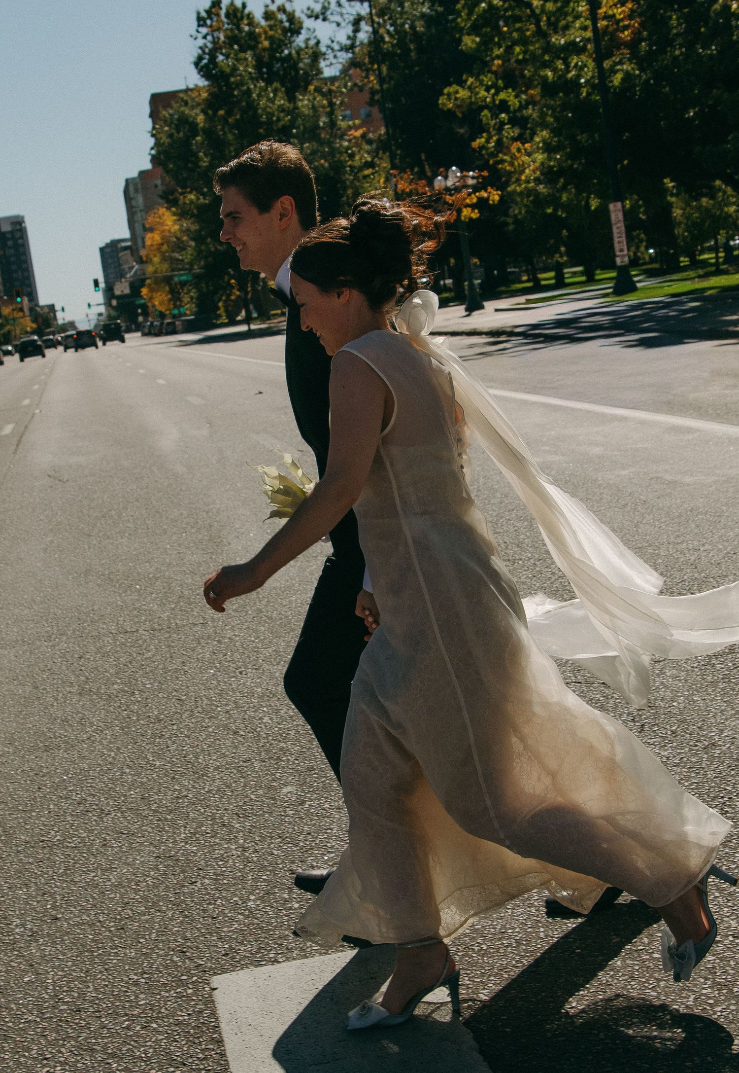 A couple runs playfully across an empty street on a sunny autumn day with trees in the background.